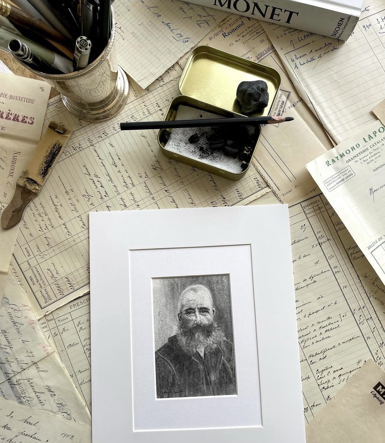 A collection of vintage documents, handwritten notes, and receipts scattered on a table. There is a framed black-and-white sketch or photograph of a bearded man in the center. On top, a small open metal box contains a piece of black charcoal or clay and a paintbrush. A list of pens is in a silver cup, and a book with the title 'MONET' is in the background.