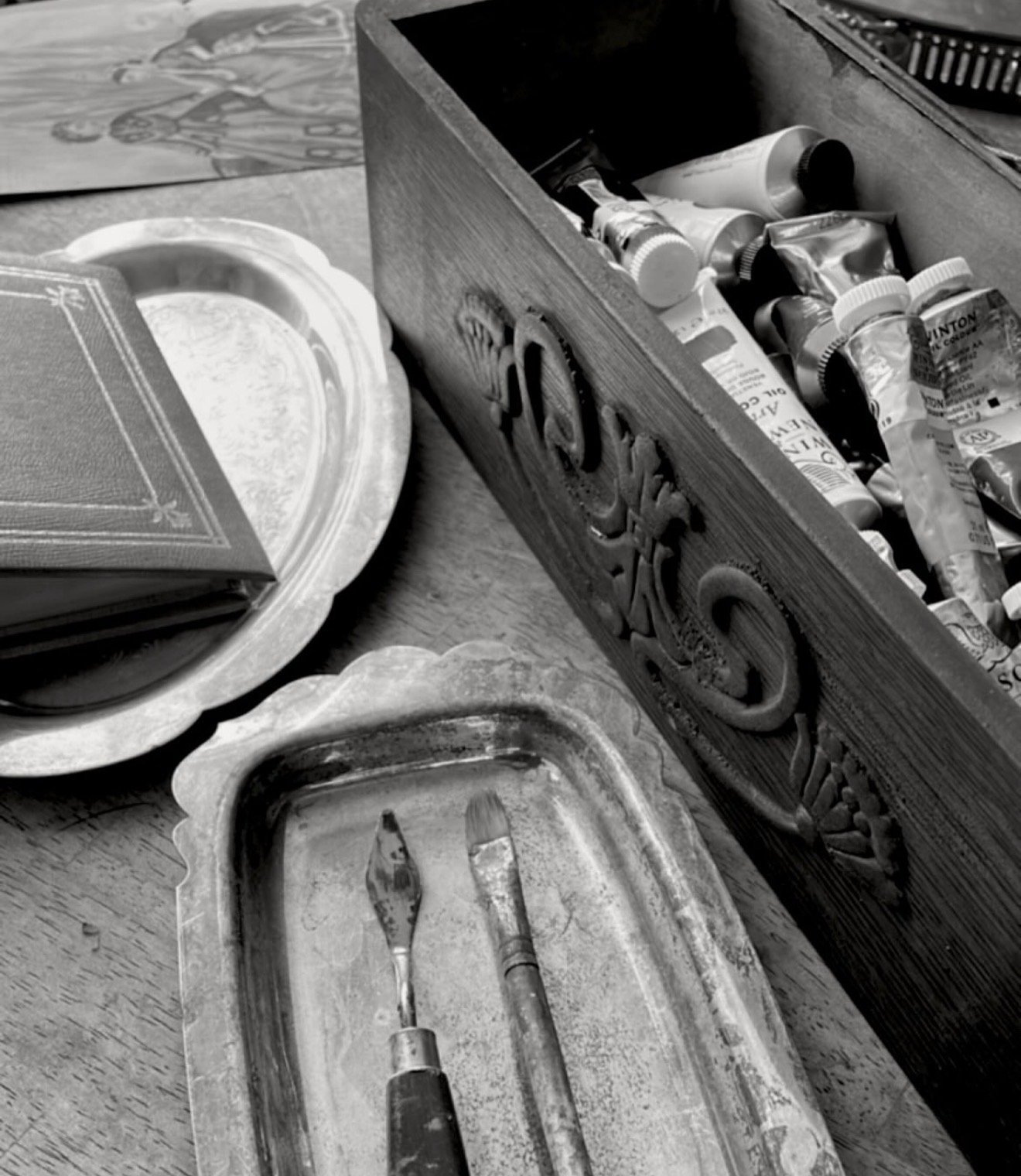 Black and white photo of an open wooden box filled with tubes of paint, a tray with two paintbrushes, a decorative book, a silver tray, and a decorated ceramic plate on a wooden table.