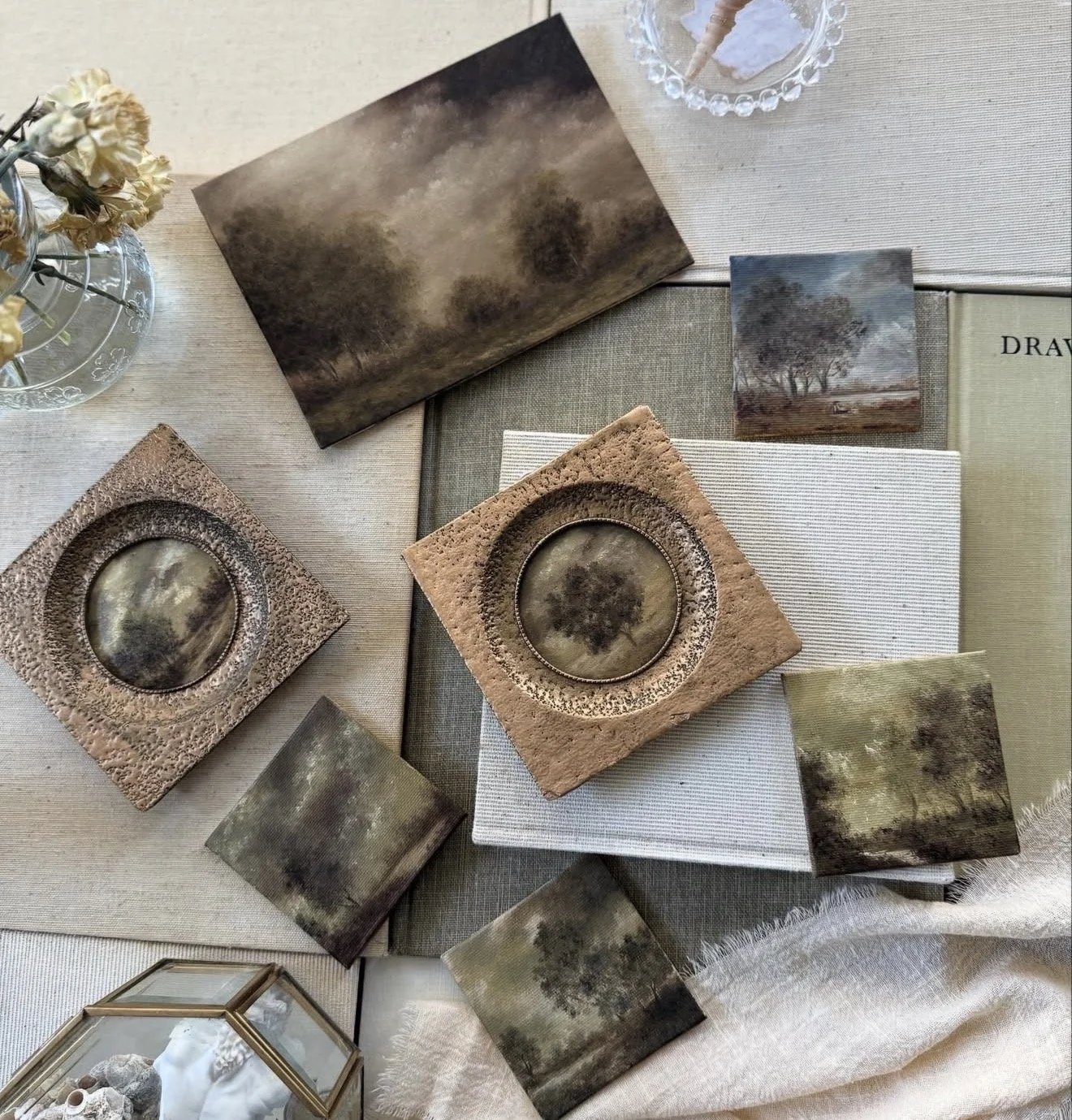 A collection of landscape-themed paintings, photo prints, and decorative items arranged on a table, including a glass vase with dried flowers, a glass dish with a snack, and books.