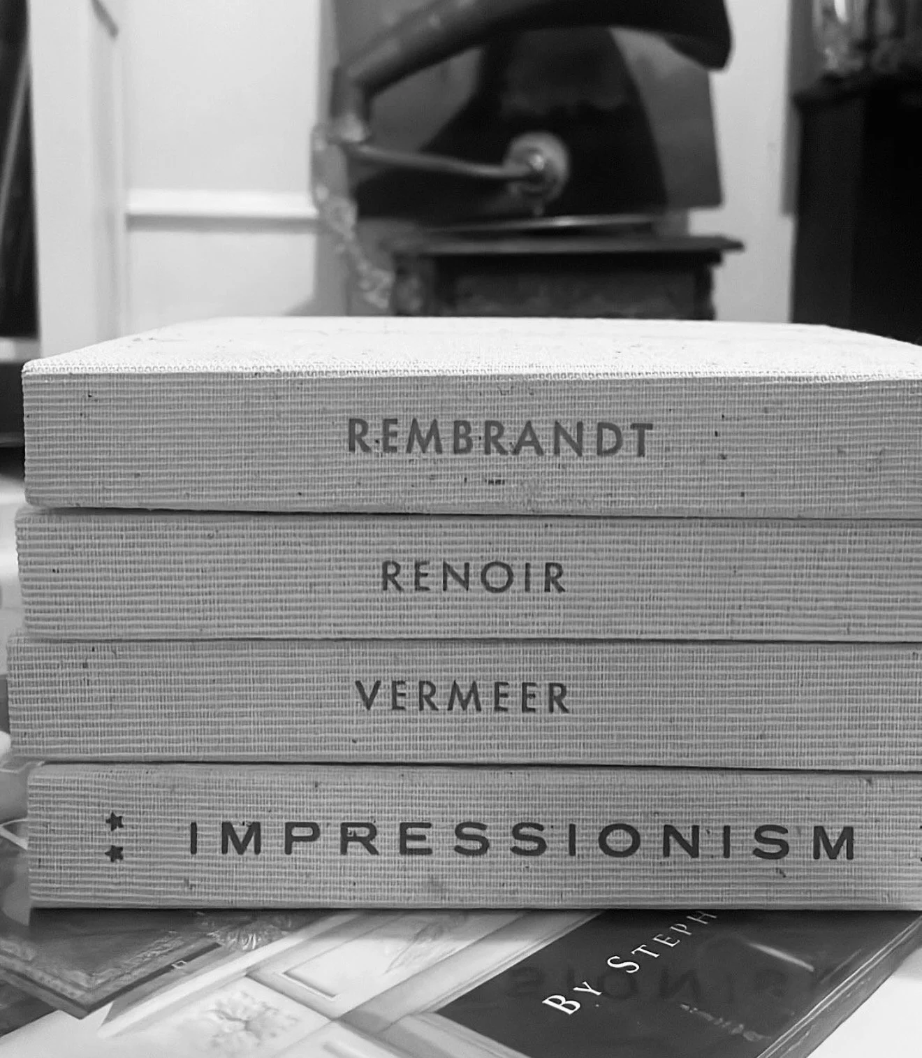 A stack of four books with textured covers, titled 'Rembrandt,' 'Renoir,' 'Vermeer,' and 'Impressionism,' with books and papers underneath in a black-and-white photo.
