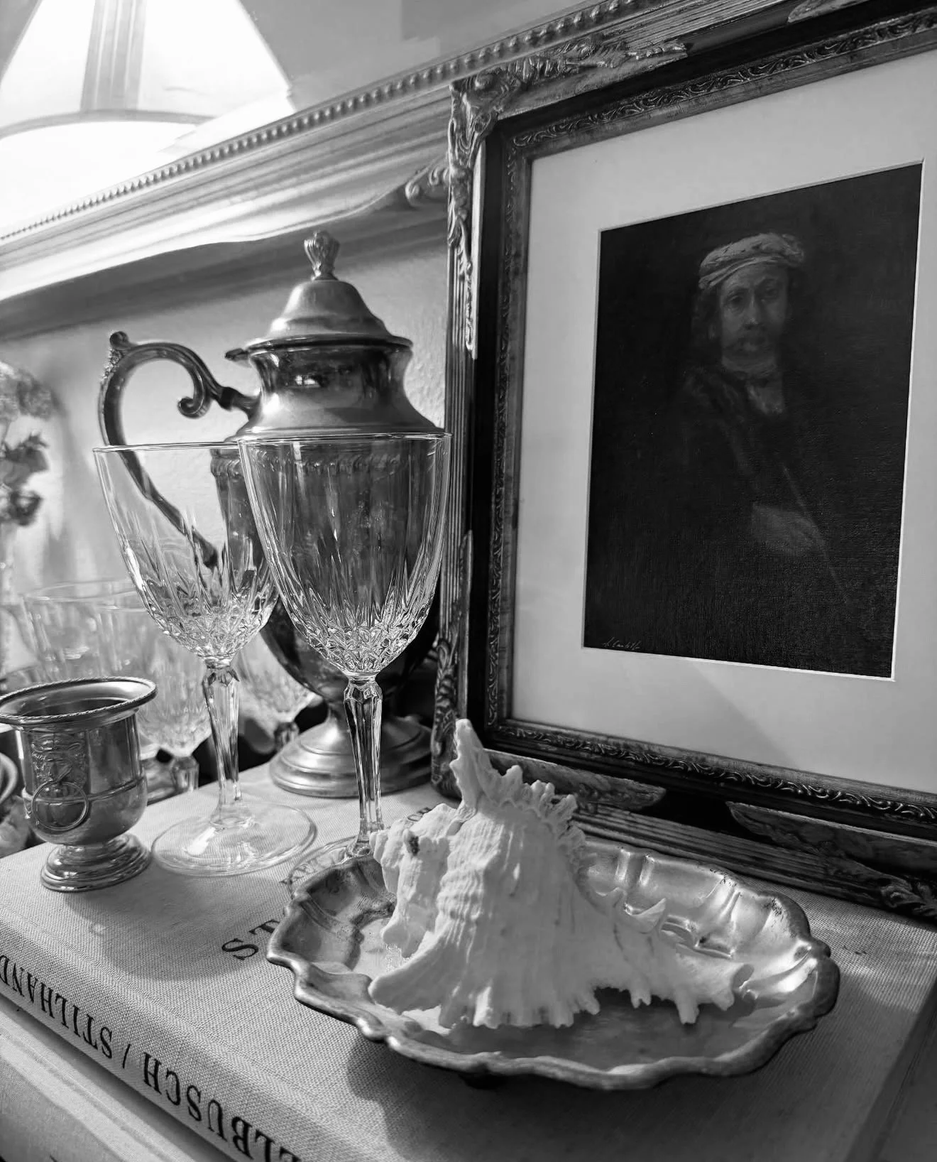Black and white photograph of a tabletop display with a framed portrait, glassware, a seashell, and a teapot, on a large book titled 'ALDEUSH / STILLANDAN'.