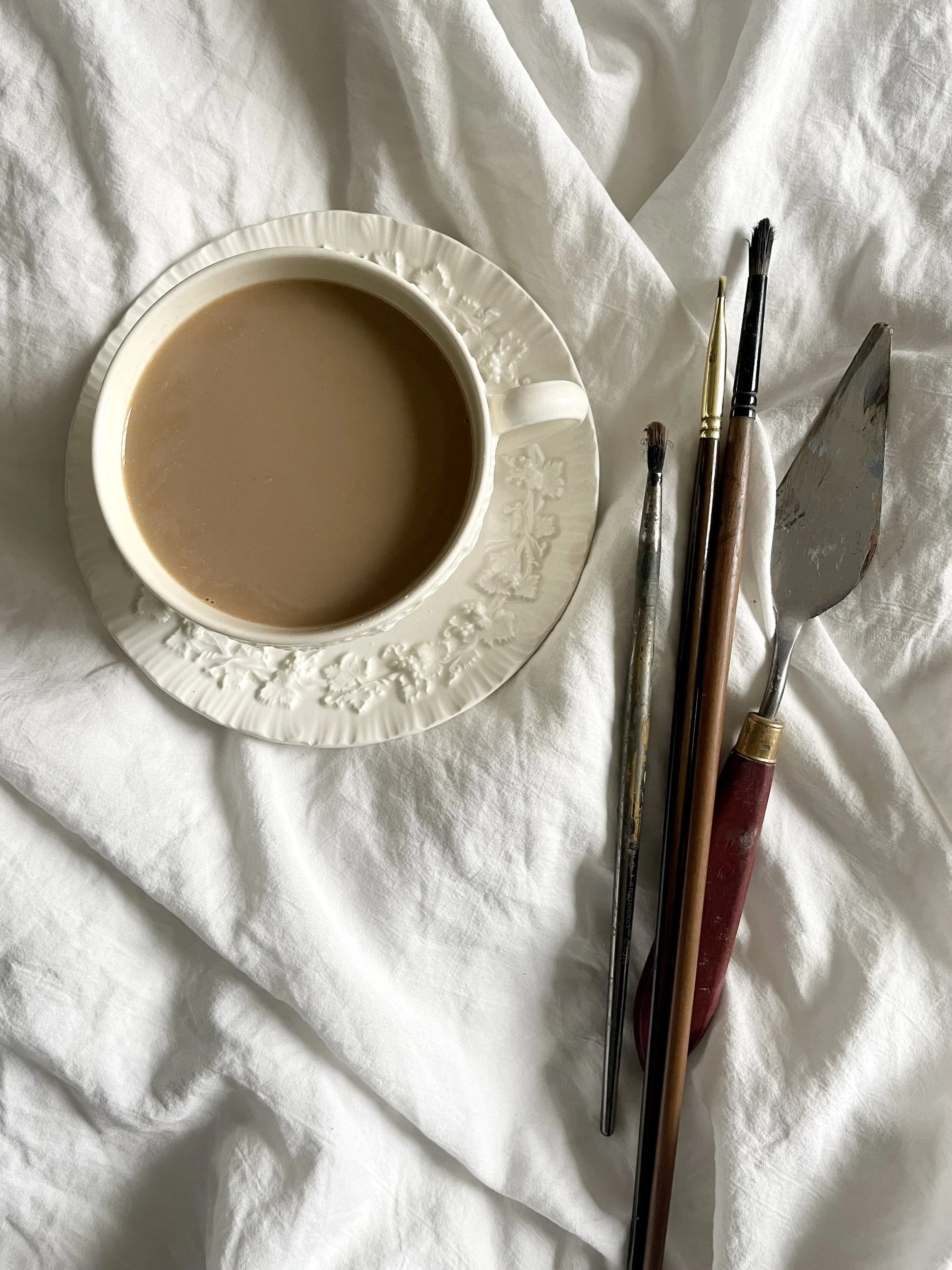 A cup of tea with milk on a white saucer, alongside paintbrushes and a small spatula on a white cloth.