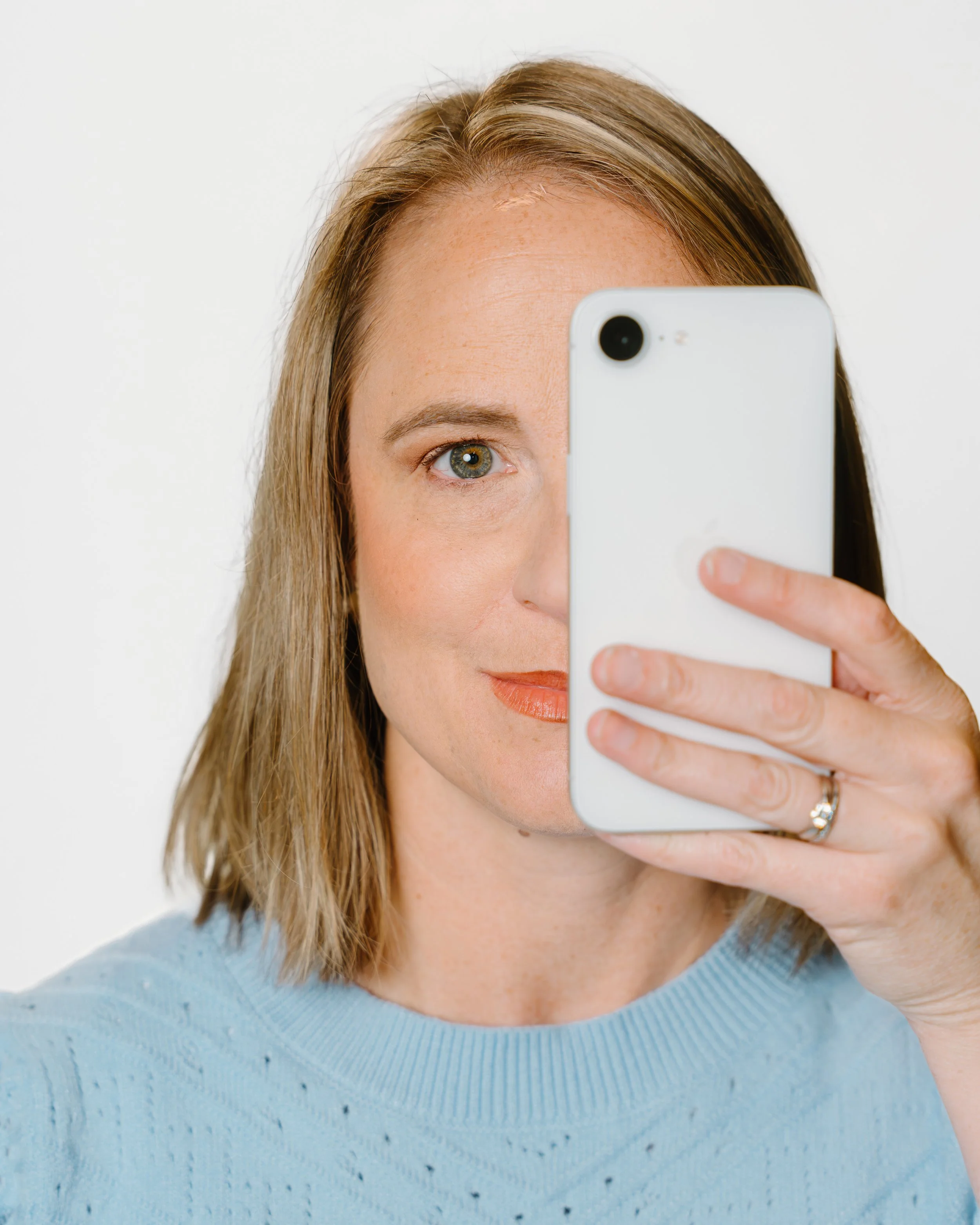 A woman with blonde hair and blue eyes taking a selfie with a smartphone, wearing a light blue sweater.