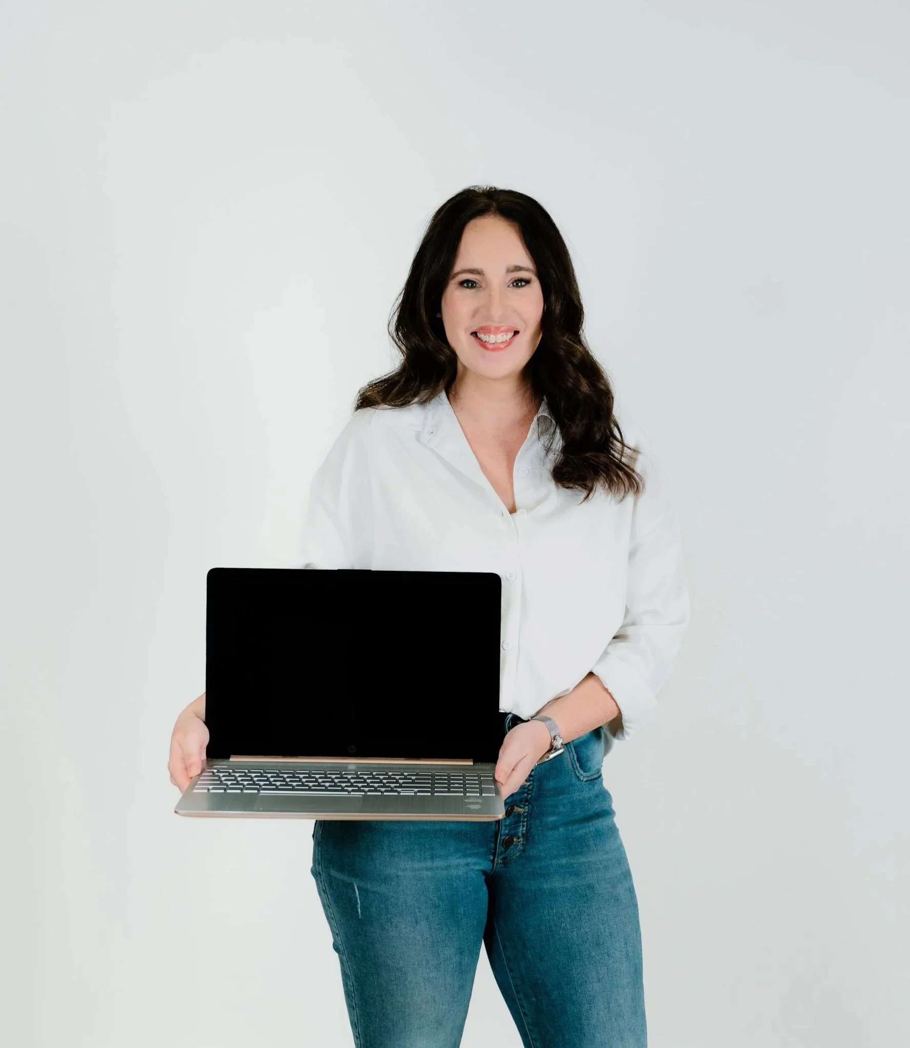 A woman with long dark hair wearing a white shirt and blue jeans, holding a silver laptop with both hands, standing against a plain white background.