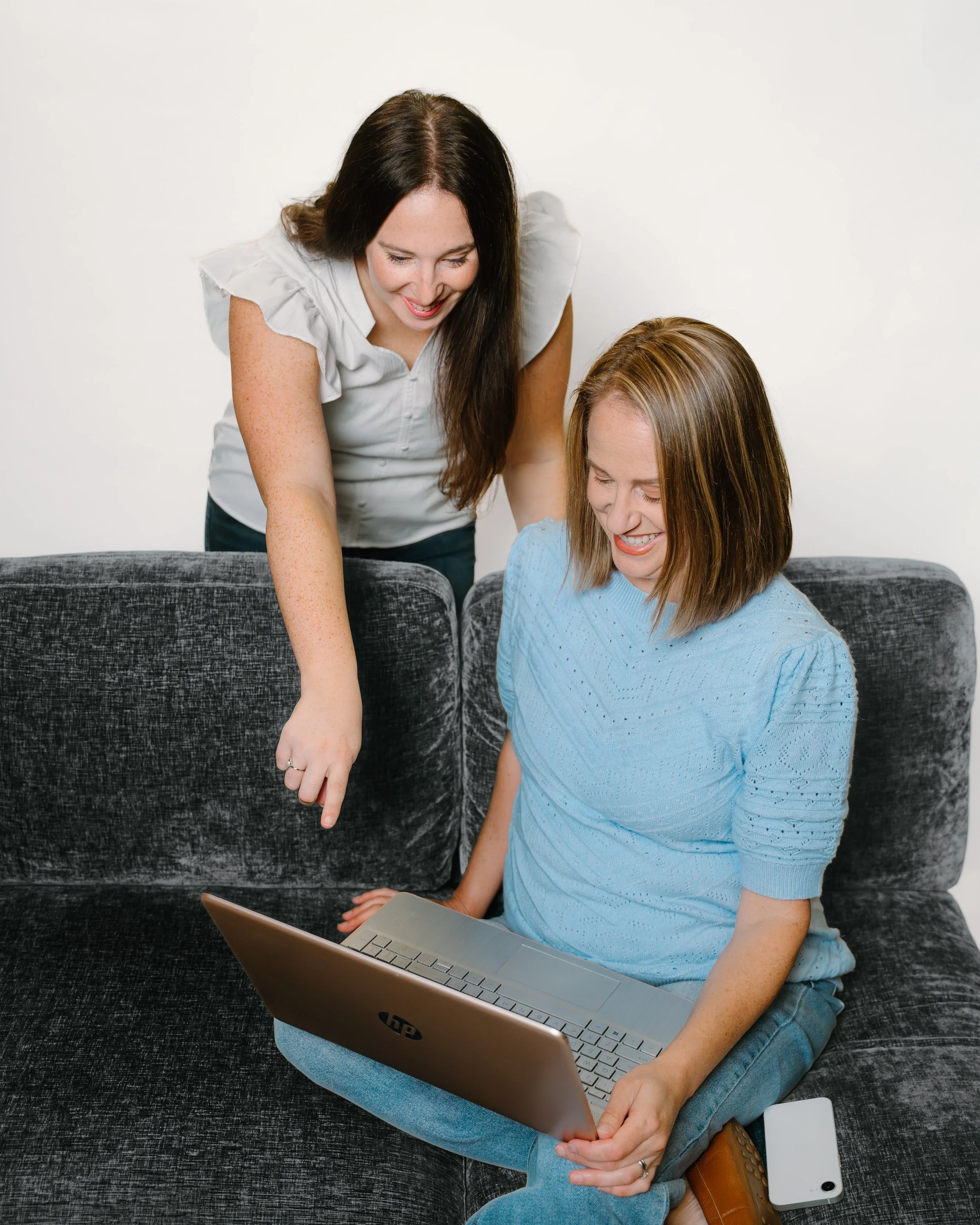 Two women, one sitting on a dark gray couch and the other standing, both smiling and looking at a silver laptop together. The woman sitting is wearing a light blue sweater and jeans, and the standing woman has long dark hair and is wearing a white blouse. There is a white smartphone and a brown purse on the couch near the seated woman.