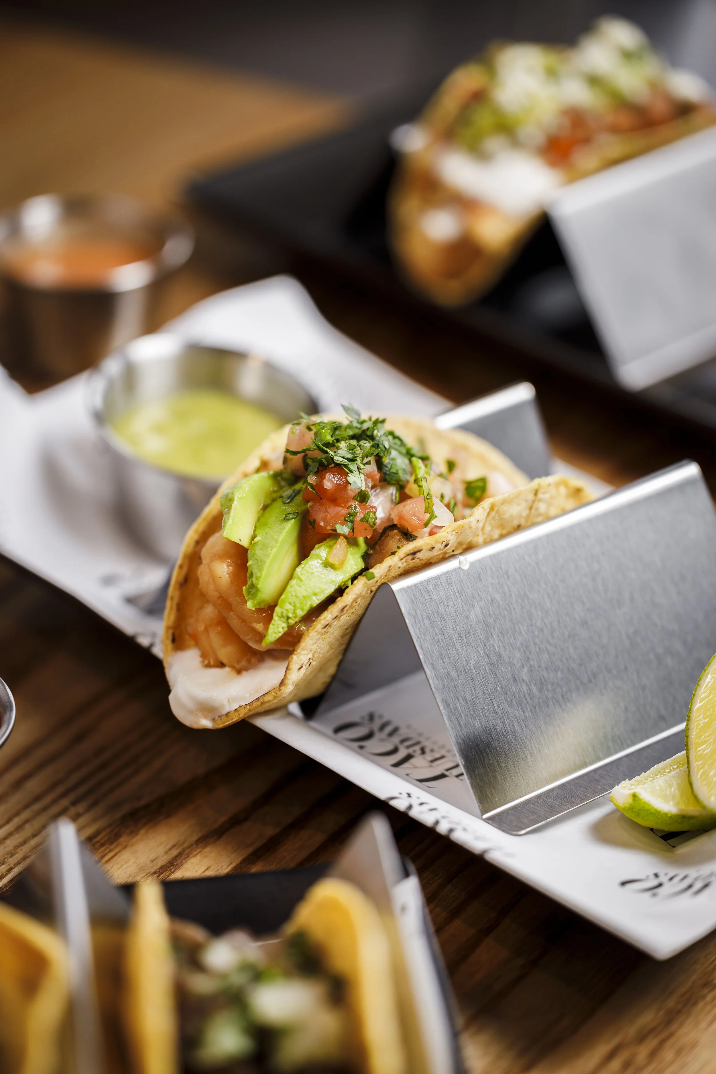 Close-up of a shrimp taco topped with avocado slices, pico de gallo, and fresh cilantro, served in a metal taco holder with a side of green sauce.