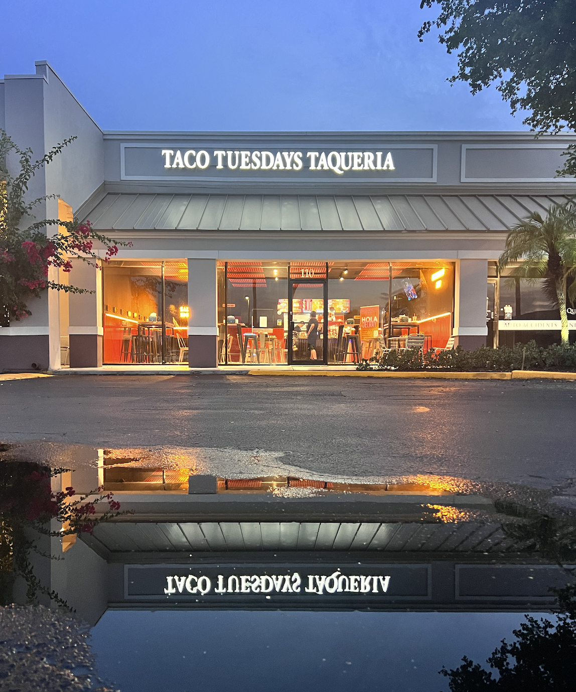 Exterior storefront of Taco Tuesdays Taqueria with illuminated restaurant sign and visible modern dining interior through glass windows at dusk