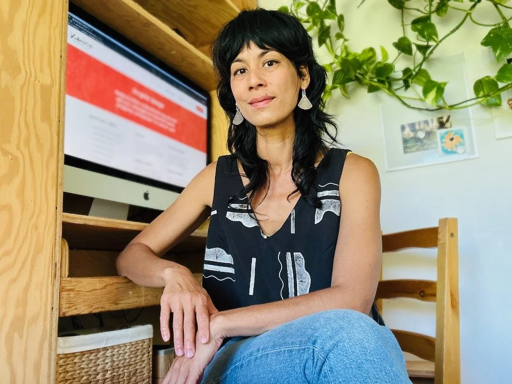 Photo of Keiko sitting at her desk, a relaxed smile, a black top and blue jeans