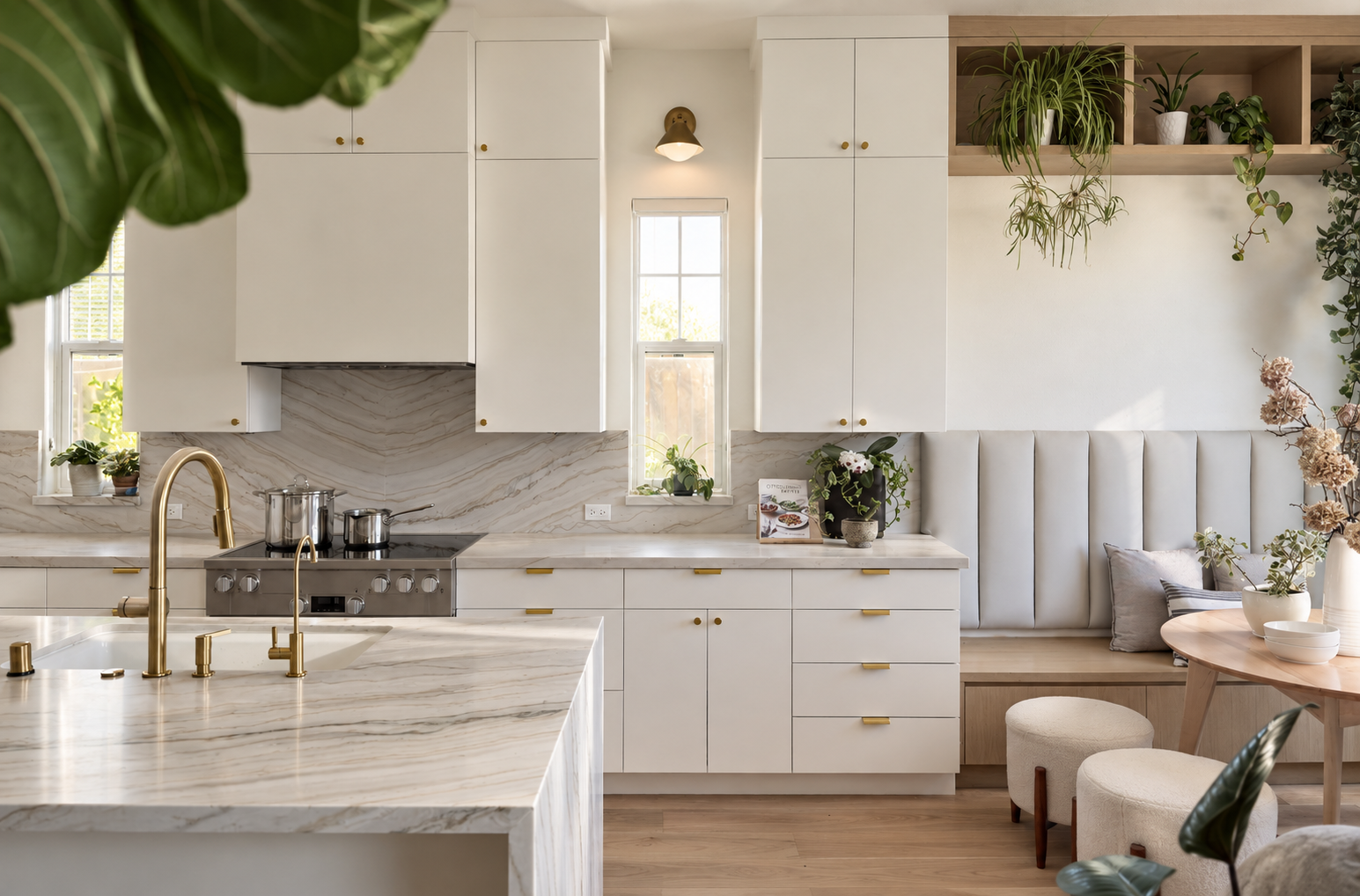 Modern kitchen with white cabinetry, marble countertops, and gold accents. There's a stove with pots, a gold faucet, and a small window with plants on the windowsill. To the right, a built-in cozy nook with a padded bench, pillows, a small round table with vases and flowers, and hanging plants on the wall.