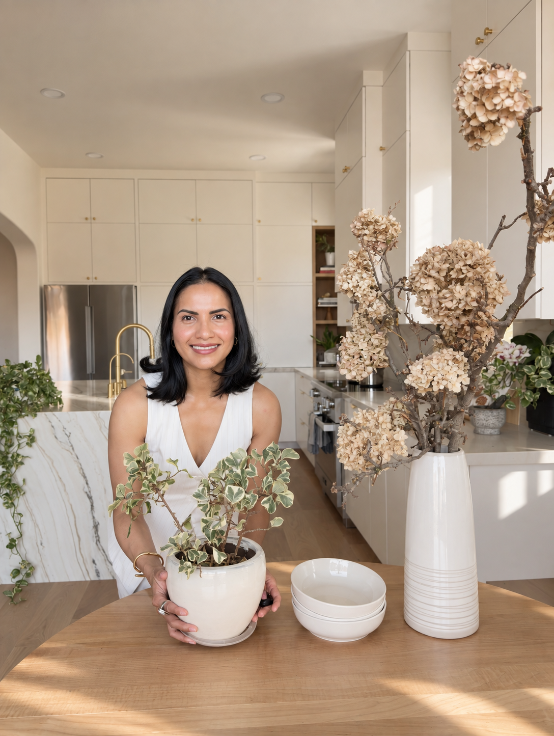 A woman in a white sleeveless top standing in a modern kitchen, placing a small potted plant with variegated leaves on a wooden table, with a large tall vase holding dried hydrangea flowers and some bowls nearby.