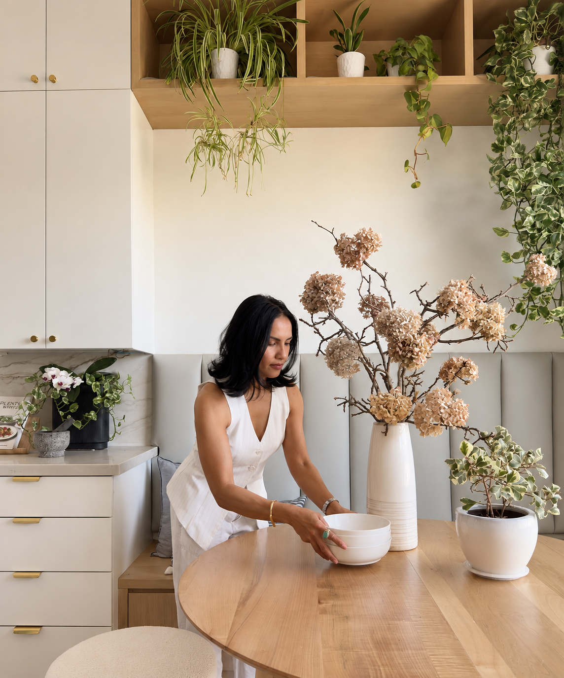 Woman arranging white bowls on a wooden dining table with large vases of dried and green plants in a bright, modern room.