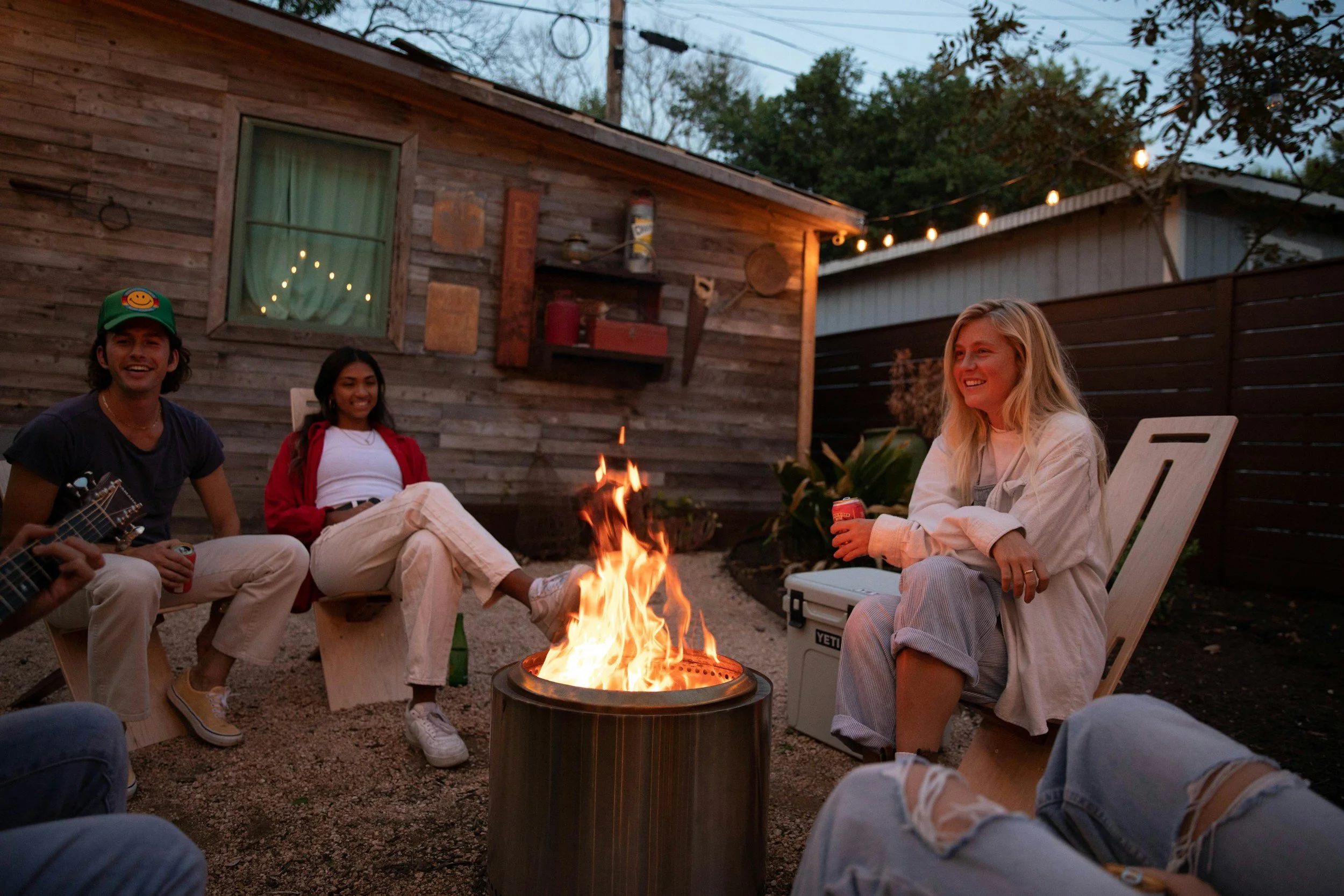 Four friends enjoying a backyard fire pit at dusk, sitting on wooden chairs and benches, with a rustic wooden house and string lights in the background.