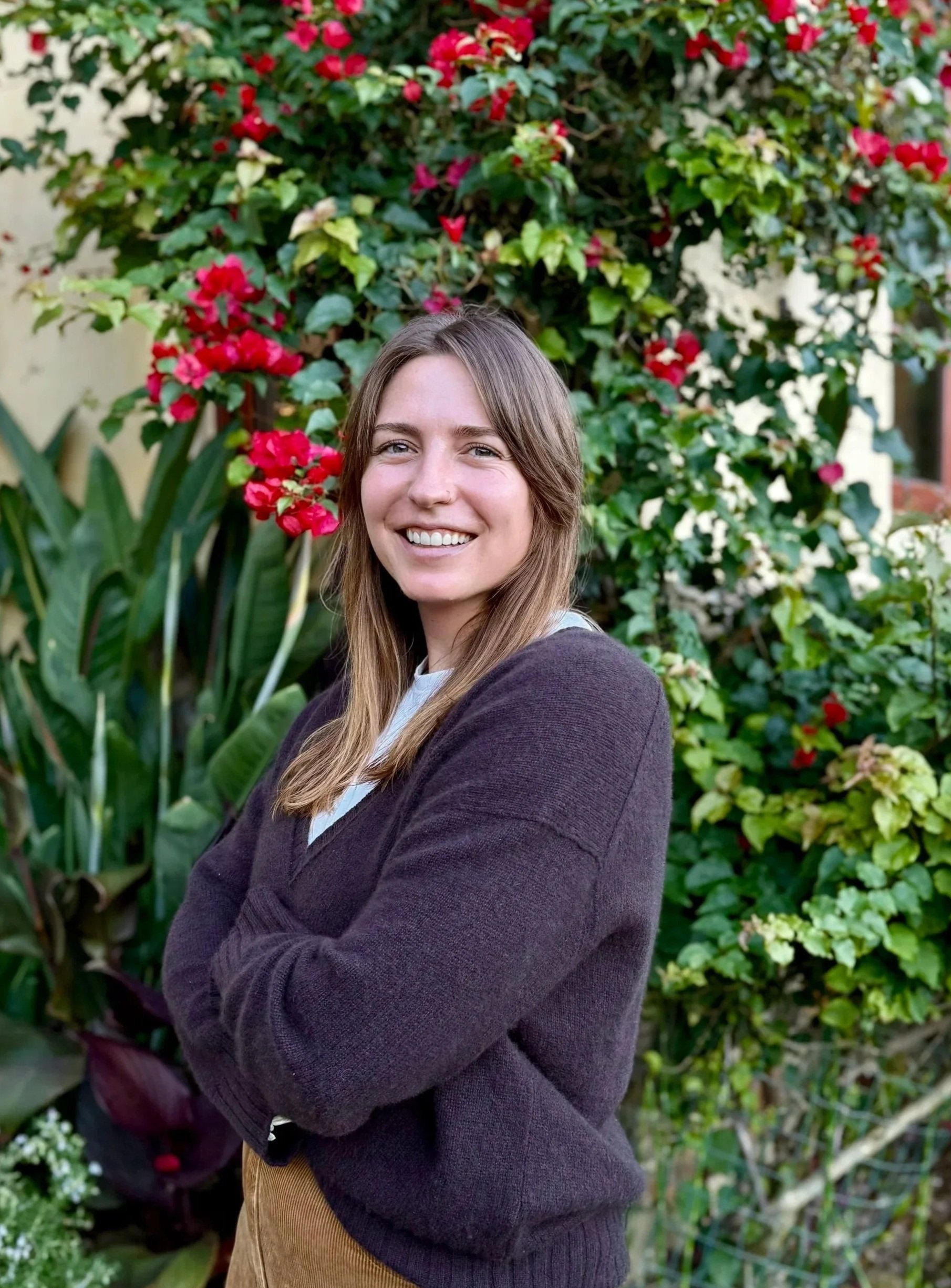 A smiling woman with long brown hair standing outdoors in front of a lush garden with red and white flowers and green plants.