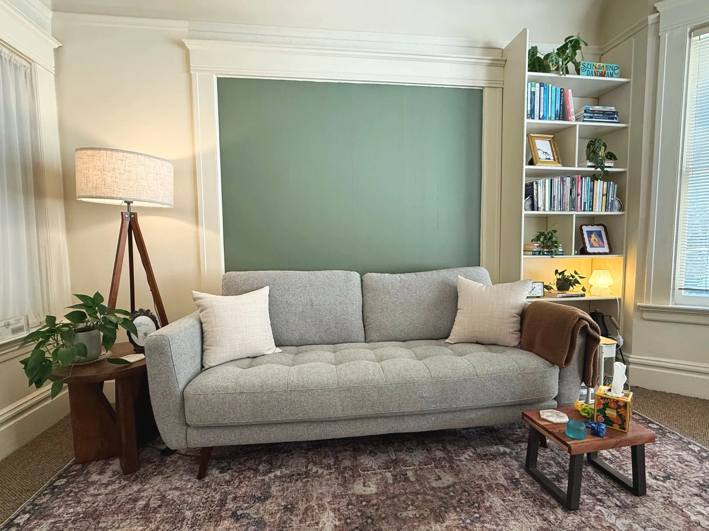 Living room with a light gray sofa, a wooden side table with a plant and a clock, a tall floor lamp, a bookcase filled with books, picture frames, and plants, a patterned area rug, and windows with blinds.