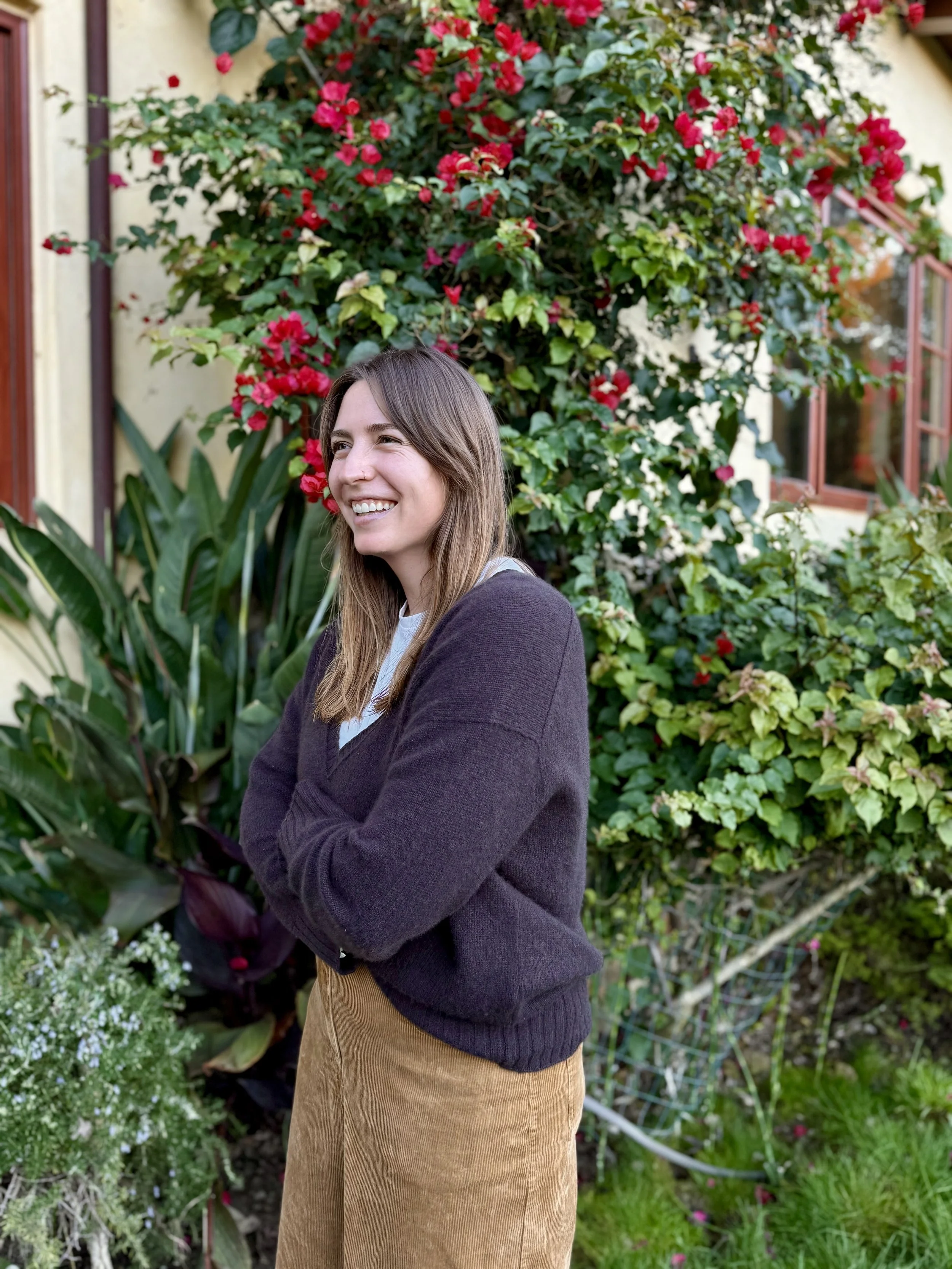 A woman with long brown hair smiling, standing with arms crossed in front of a garden with green plants and bright red flowers, near a house with yellow walls and red window frames.
