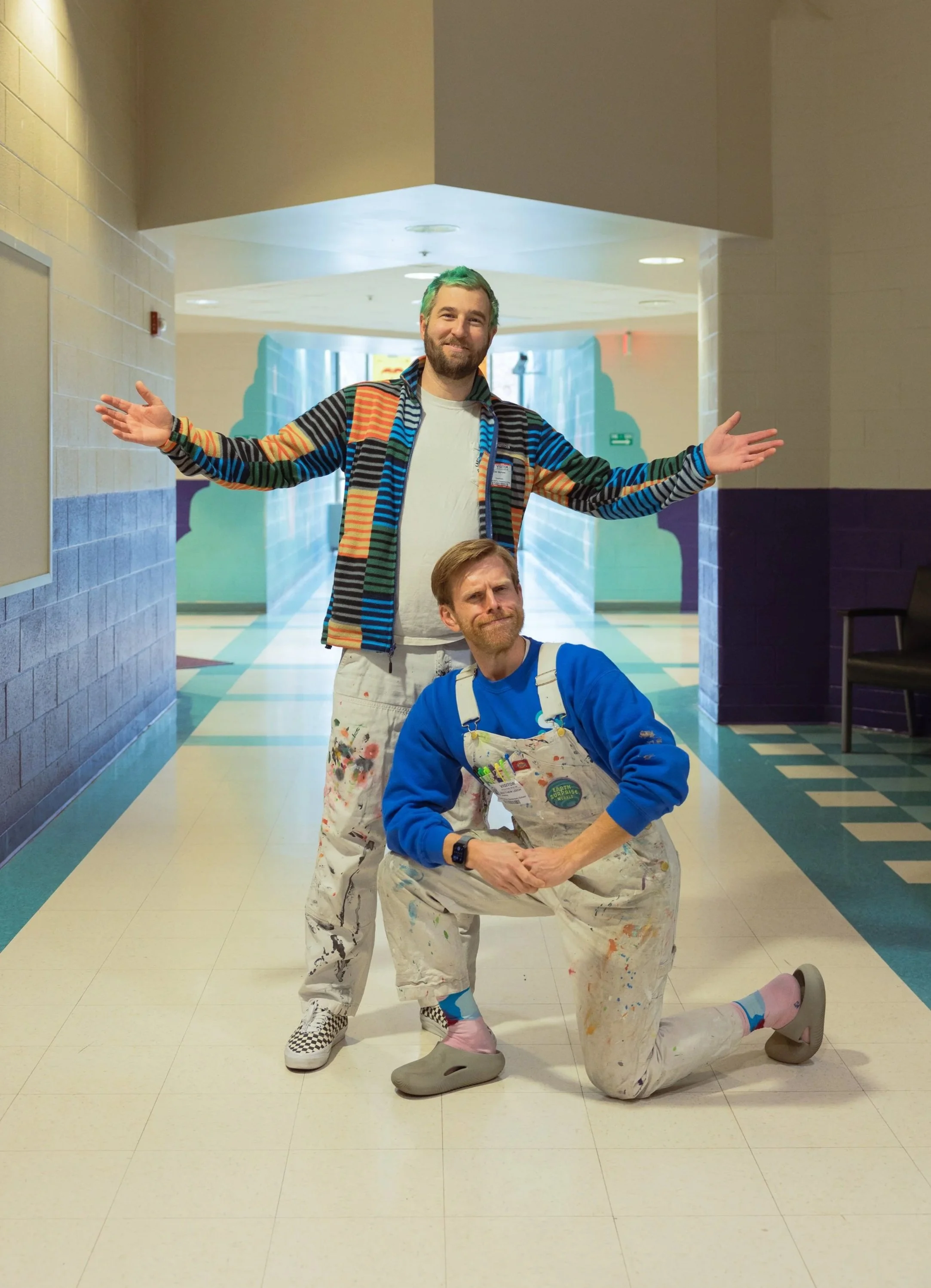 Two men with painted hair and colorful outfits posing in a school hallway. One man is standing with arms outstretched, and the other is kneeling with a hand on their thigh. Both have smiles and paint splatters on their clothes.