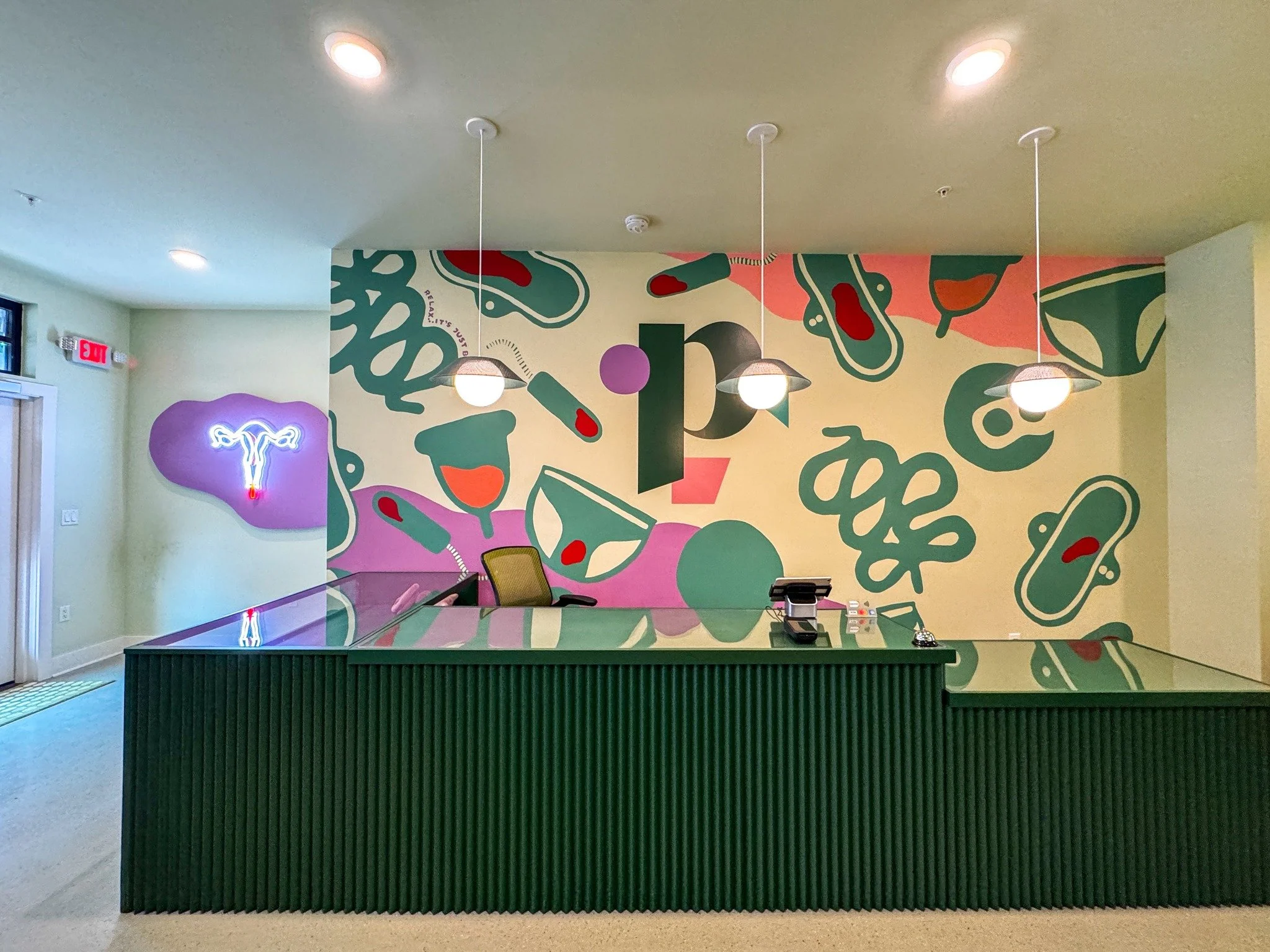 A modern reception desk with a dark green front panel, a green marble countertop, and colorful abstract wall art in the background featuring various shapes and patterns. There are ceiling lights and a neon sign of a uterus on the wall.