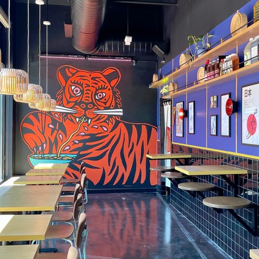Interior of a restaurant featuring a large, vibrant mural of an orange tiger eating noodles on a black wall. The space has wooden tables, chairs, and decorative hanging lights.
