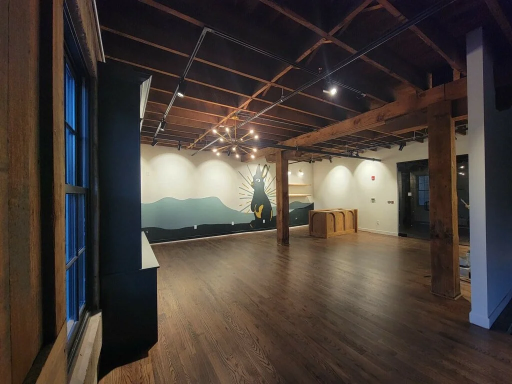 Empty interior space with wooden floors, exposed ceiling beams, and a large mural of a mountain and a playful black creature on the wall.