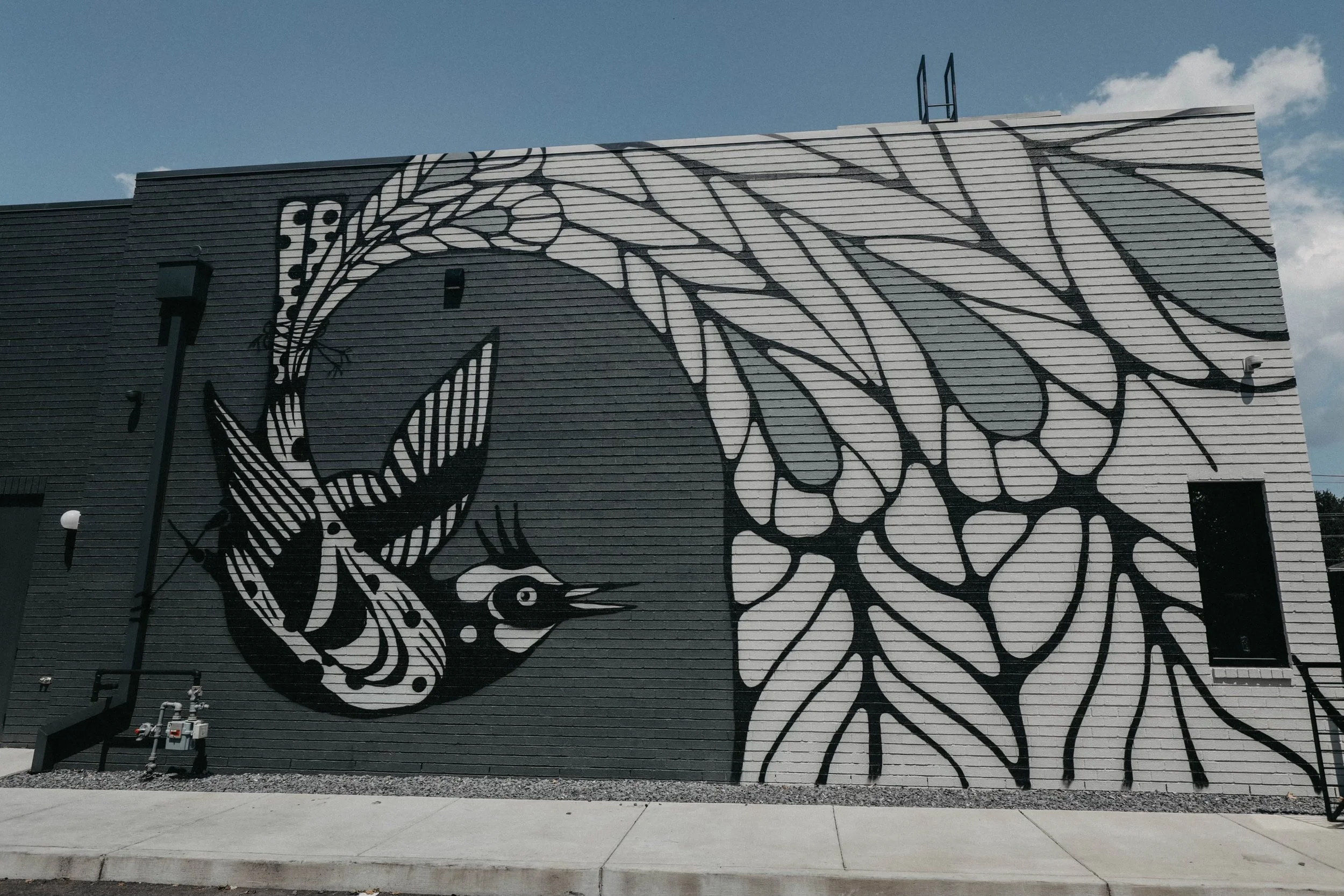 Black and white mural of a bird with leaf-like patterns on its wings painted on a building's brick wall.