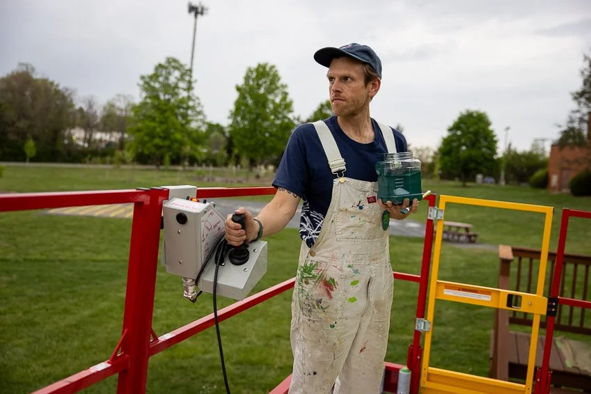 A man in a blue cap and navy t-shirt, wearing white paint-splattered overalls, stands on a red lift platform outdoors holding a container of blue paint in one hand and a paint sprayer in the other, with trees and a park in the background.