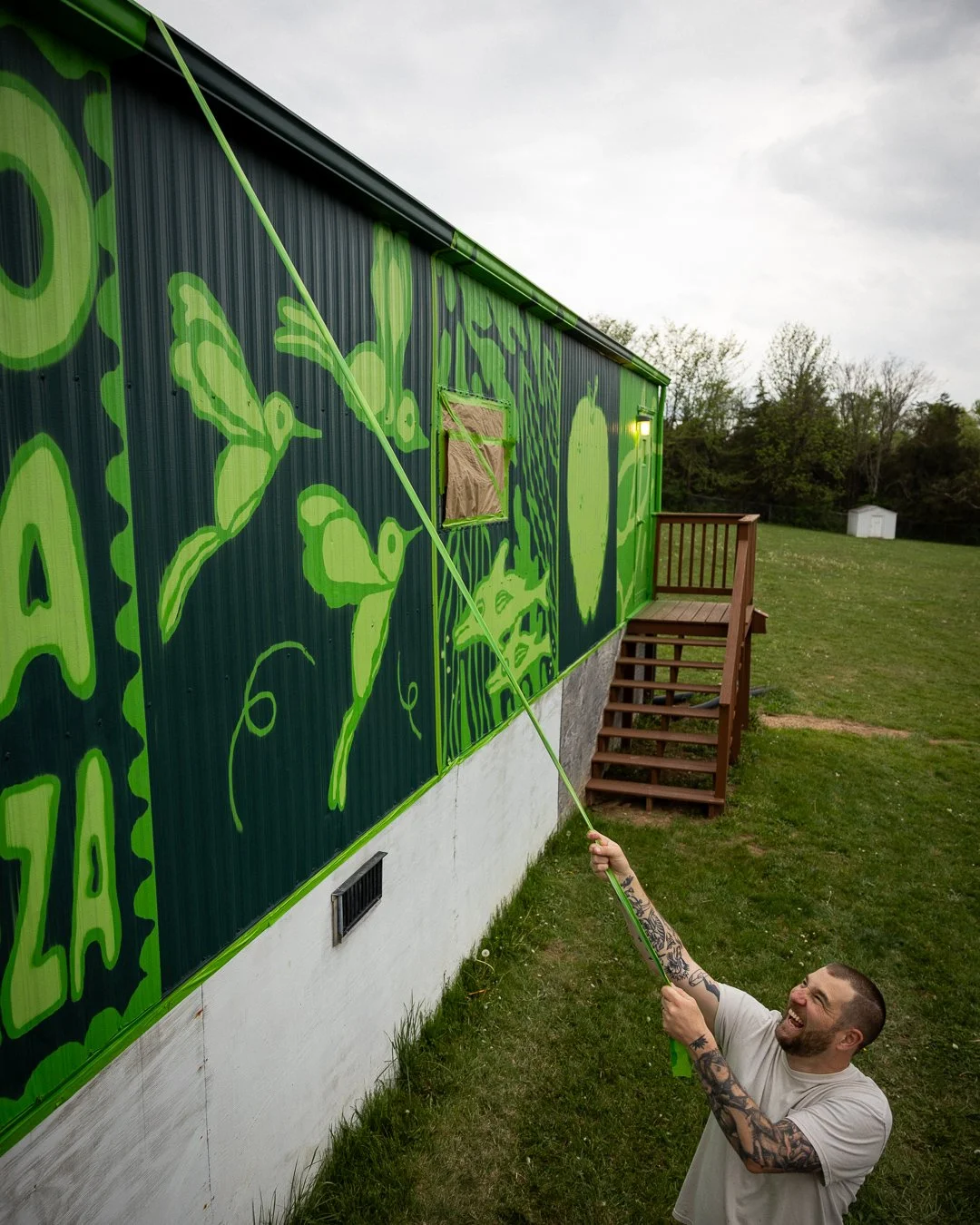 A man with tattoos on his arms is pulling a green rope to paint a mural of vibrant green abstract shapes on the side of a house with dark siding, with a small window covered by a brown paper