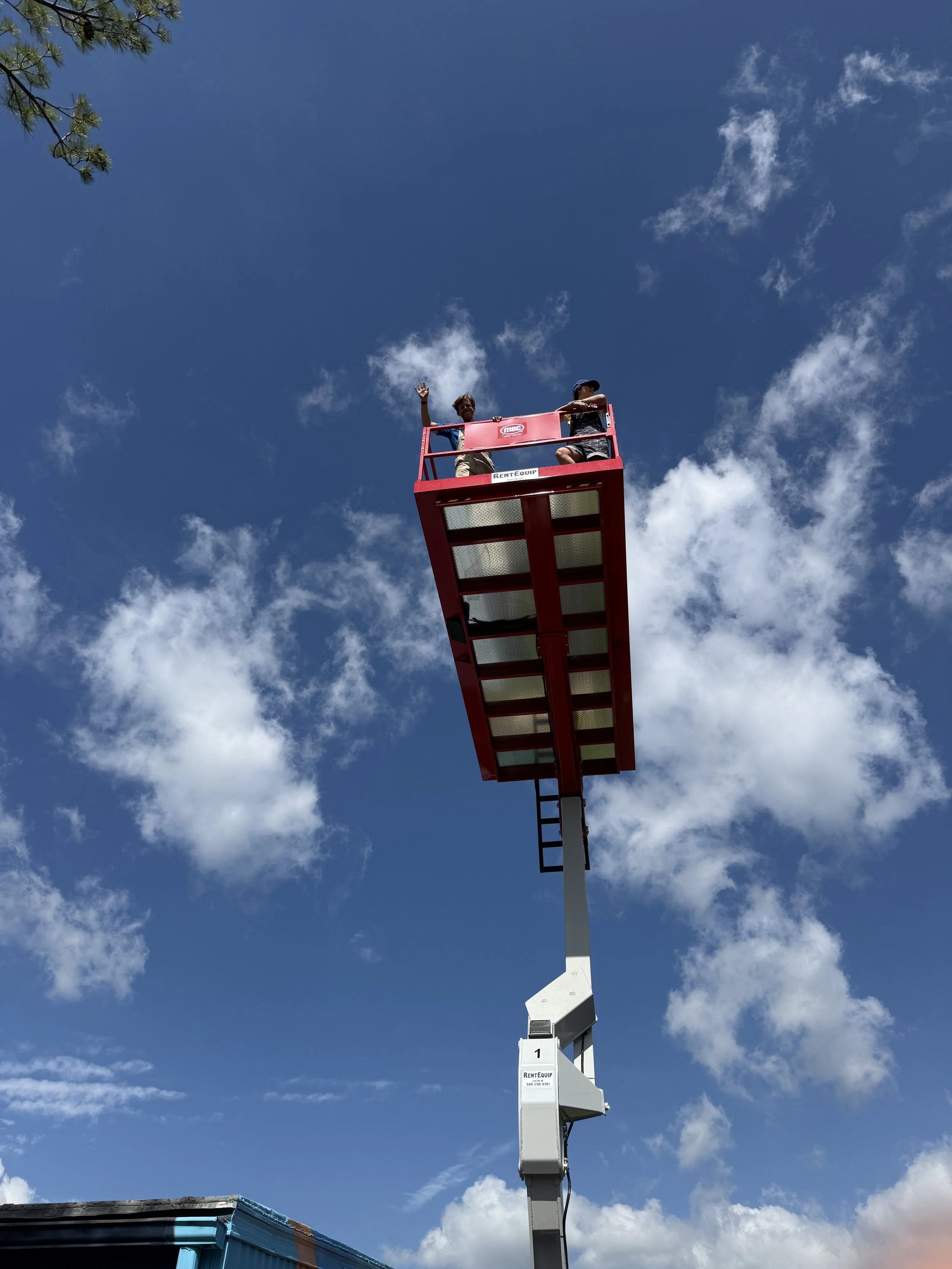 Two children on a red cherry picker lift, waving and looking down at the camera against a partly cloudy blue sky.