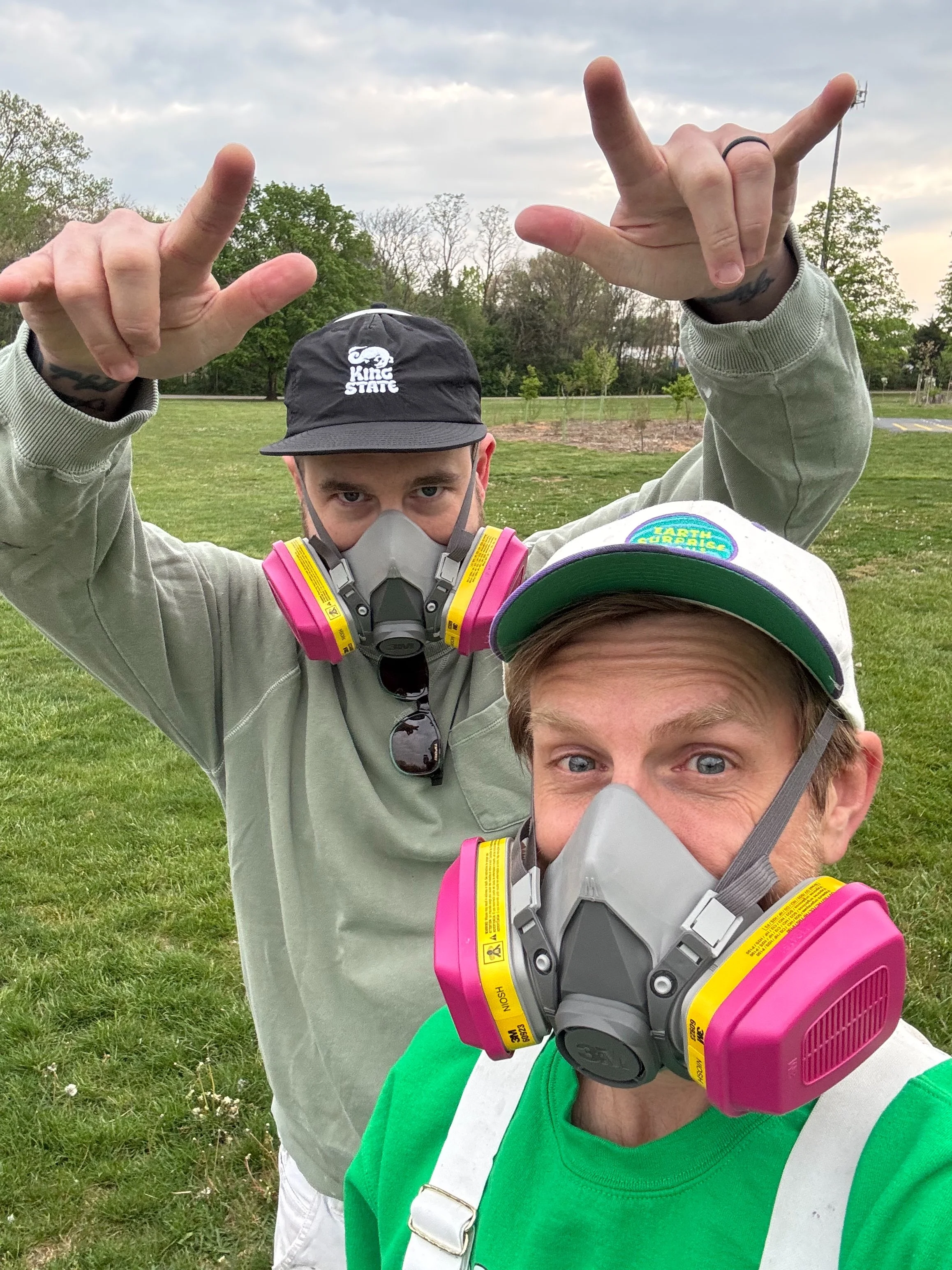 Two men stand outdoors in a grassy area, both wearing safety masks with pink filters and making hand signs. One man wears a gray hoodie and a black cap, while the other wears a green and white shirt and a white cap with a colorful patch. Trees and cloudy sky are in the background.