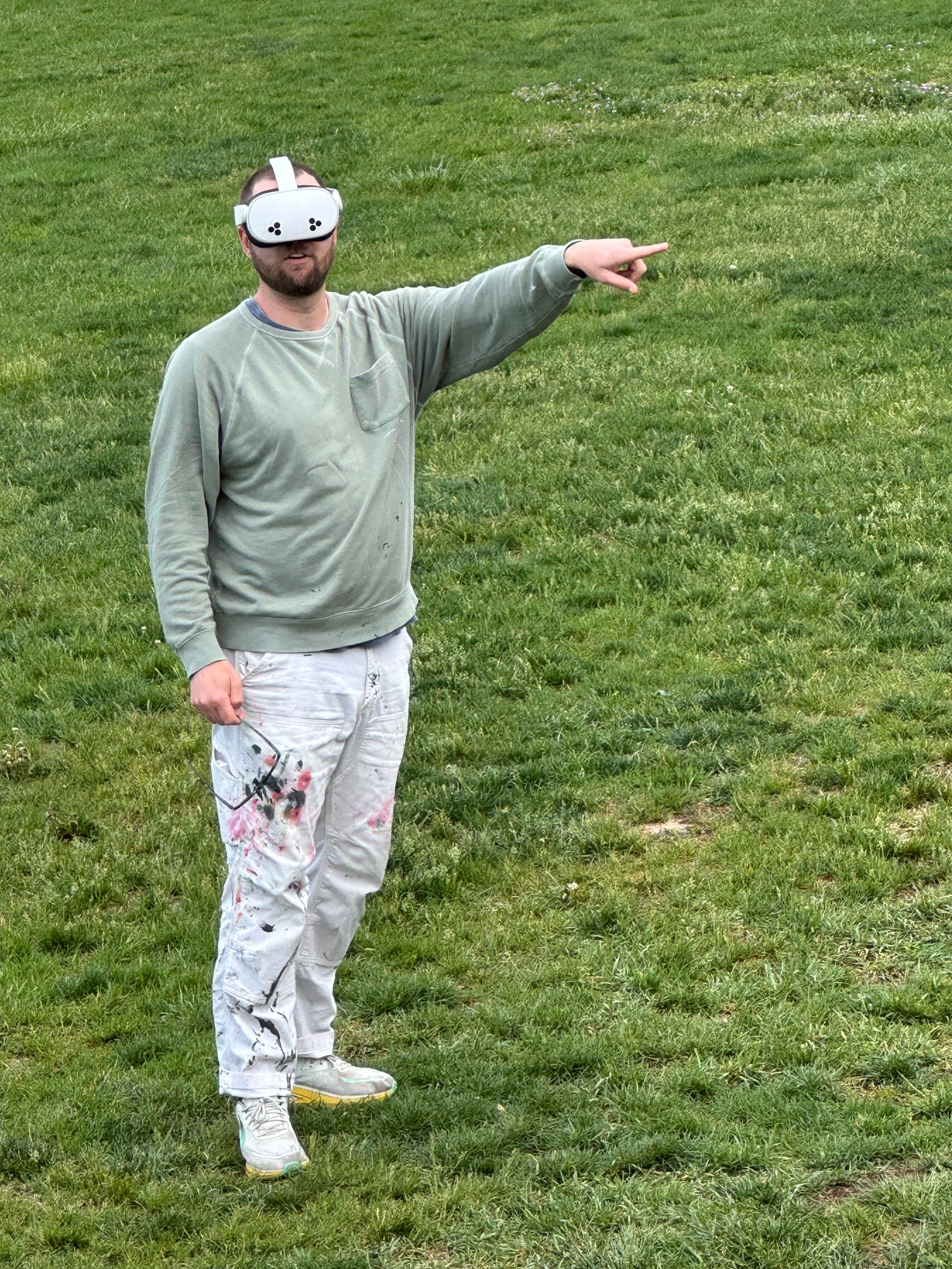 A man wearing a virtual reality headset standing on grass, pointing with his right hand, and dressed in paint-splattered white pants and sneakers.