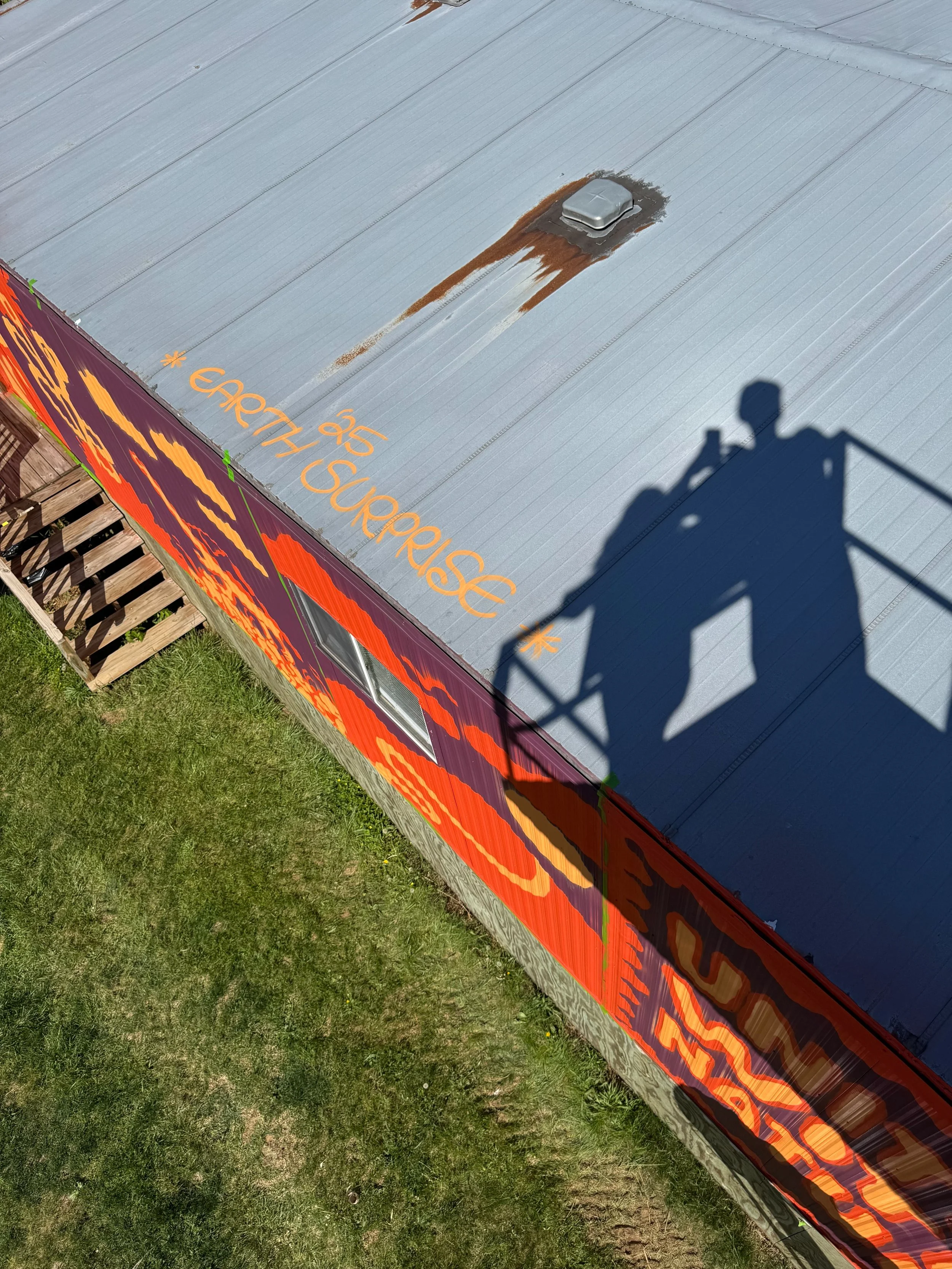 Image shows a rooftop with a chimney and some signs of rust around the chimney. There is colorful signage along the side of the building and a wooden staircase at the bottom left. The shadow of a person on a lift is cast on the roof.