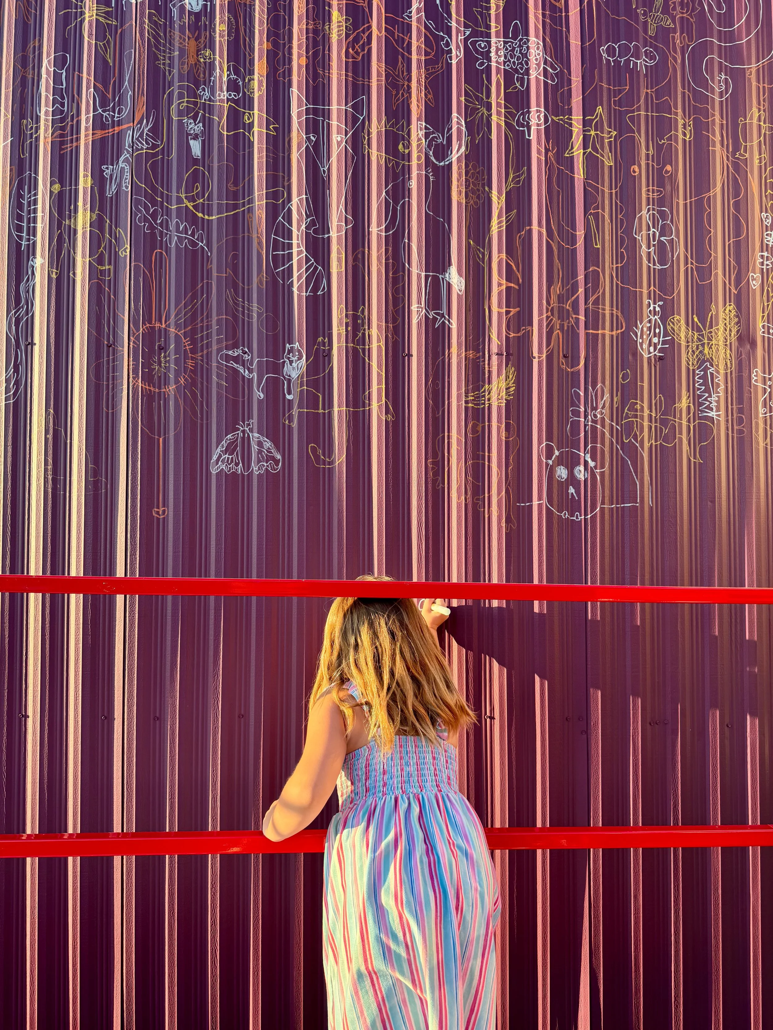 A young girl with long hair wearing a colorful striped dress is standing in front of a purple wall decorated with chalk drawings of animals, stars, and flowers. She is reaching up with her right arm.