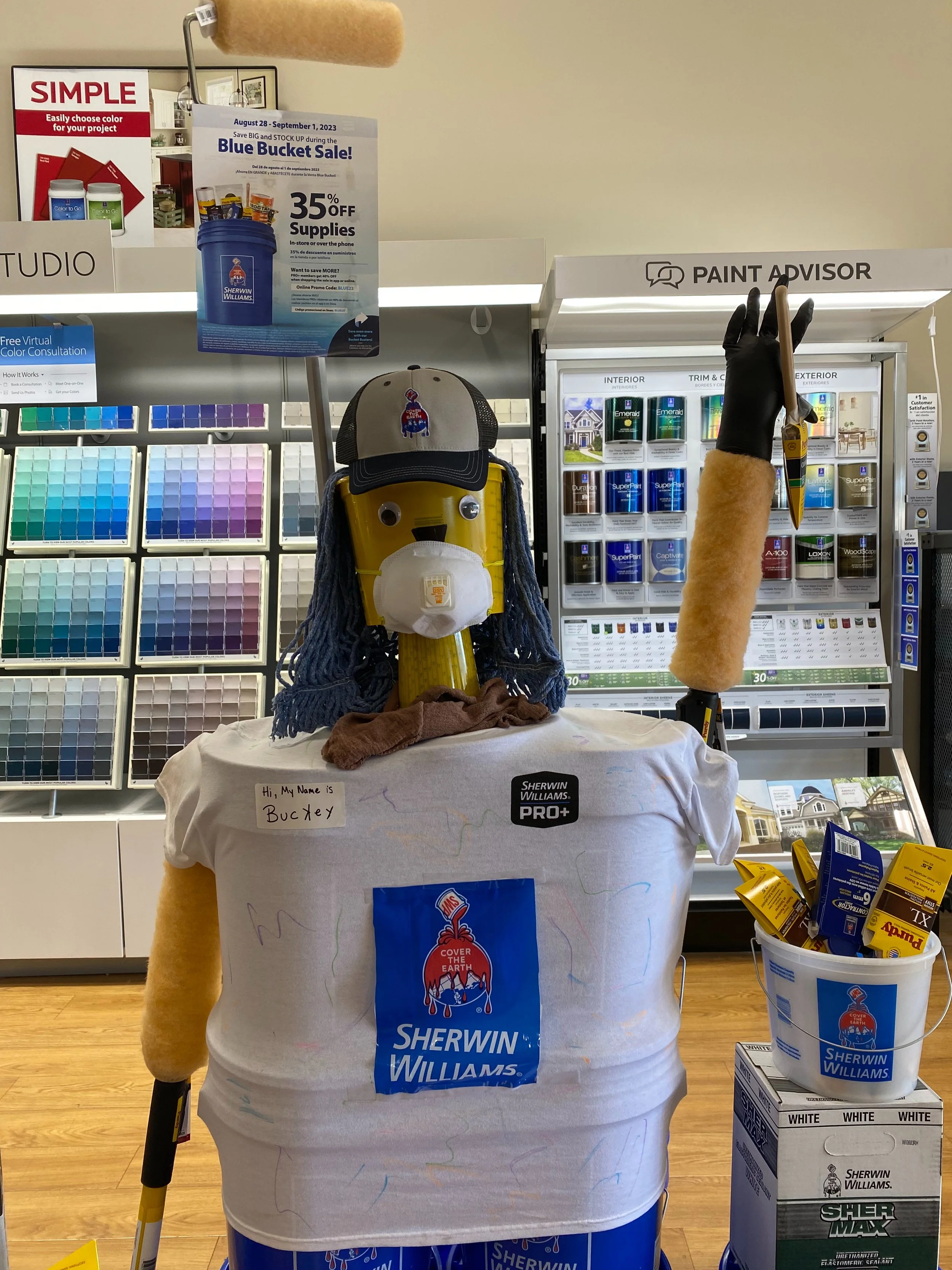 Display of Sherwin Williams paint sample cards, a yellow and black cane, and a mannequin with a white t-shirt that has a drawing and a name tag. The setup appears in a home improvement store, with various paint cans and supplies visible in the background.