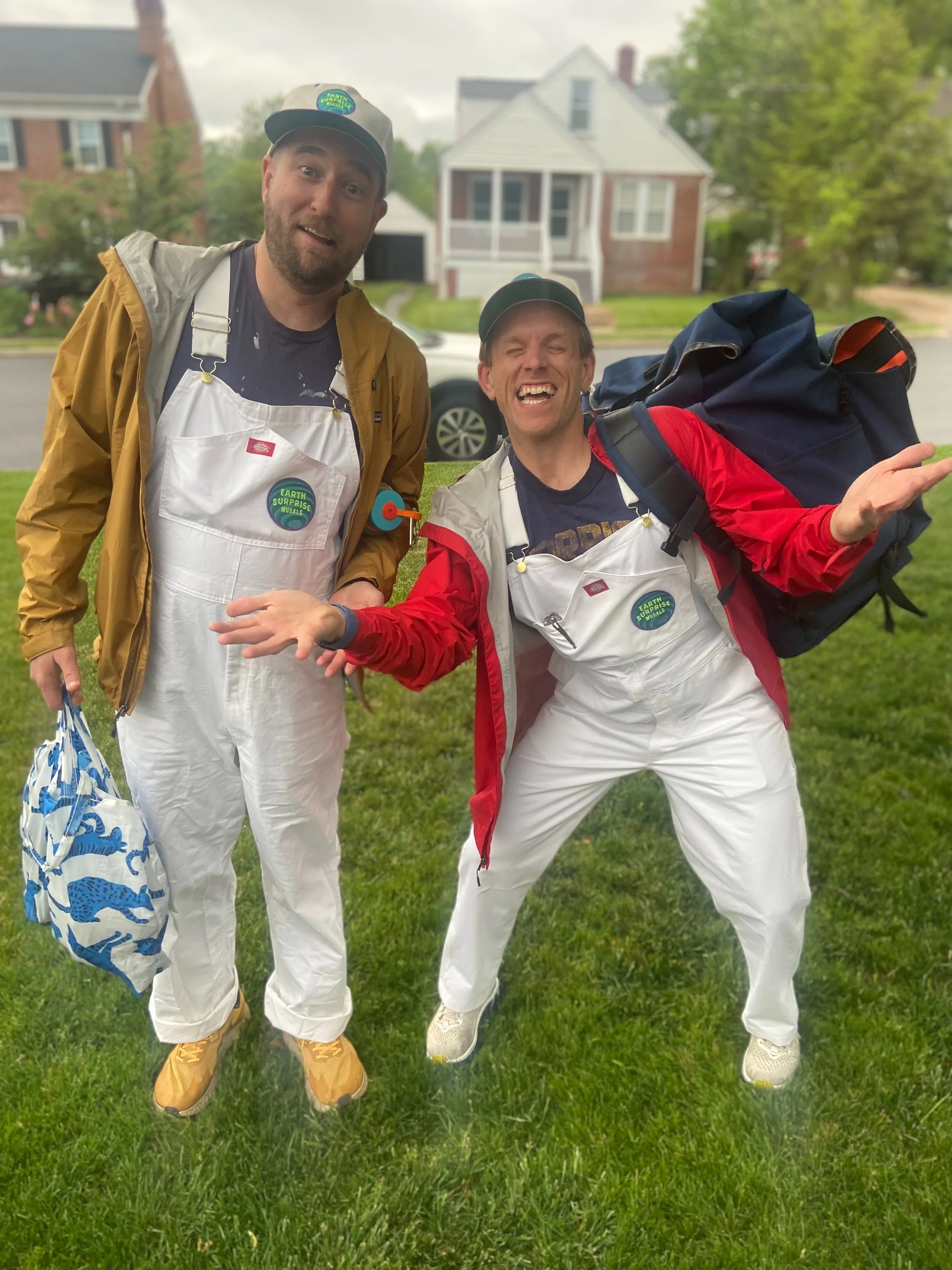 Two men smiling and making playful gestures in a grassy front yard with houses in the background. Both are wearing white overalls with patches that say "Earth Surprise Murals," one in a red jacket and carrying a large backpack, the other in a tan jacket with yellow shoes.