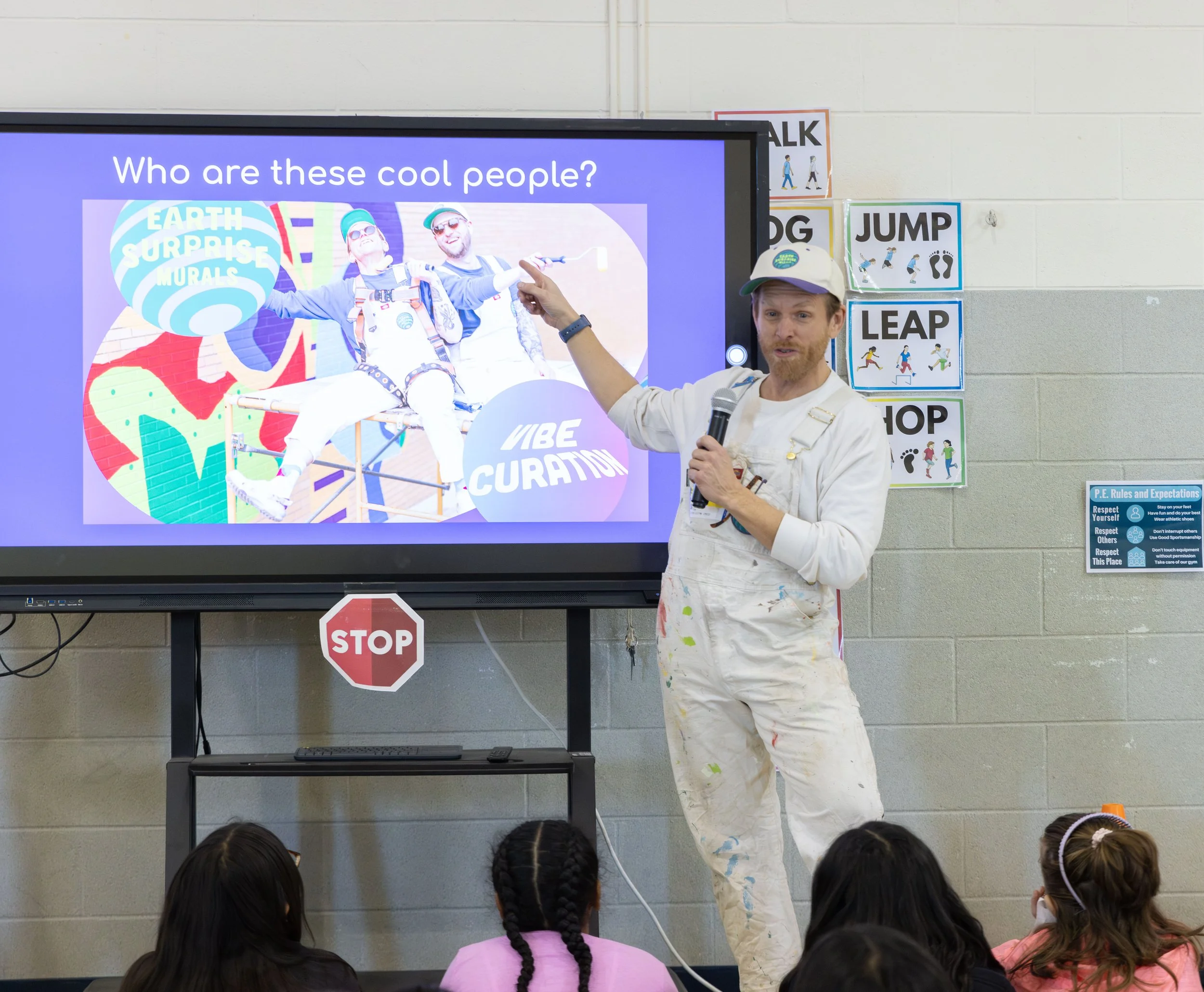 A man with a beard and red hair wearing white overalls, holding a microphone and pointing at a large screen, appears to be giving a presentation to young children. The screen displays a colorful image of two people sitting on a bench with the text 'Who are these cool people?' The classroom wall behind them features signs with words like 'TALK,' 'JUMP,' and 'LEAP,' and a smaller sign with 'P.E. Rules and Expectations.'