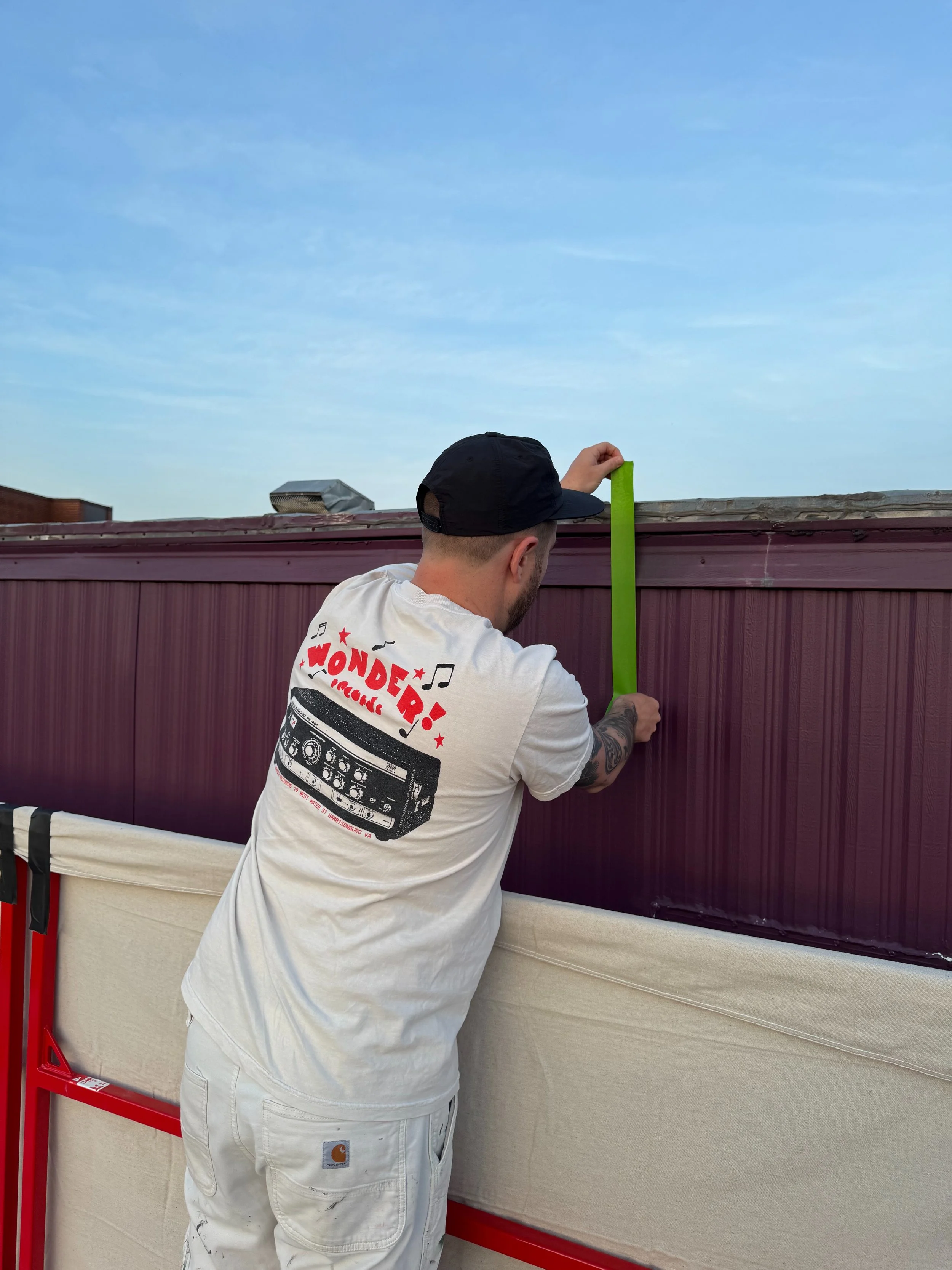 Person measuring a fence with a green measuring tape outdoors, wearing a cap and a white t-shirt with a DJ graphic.