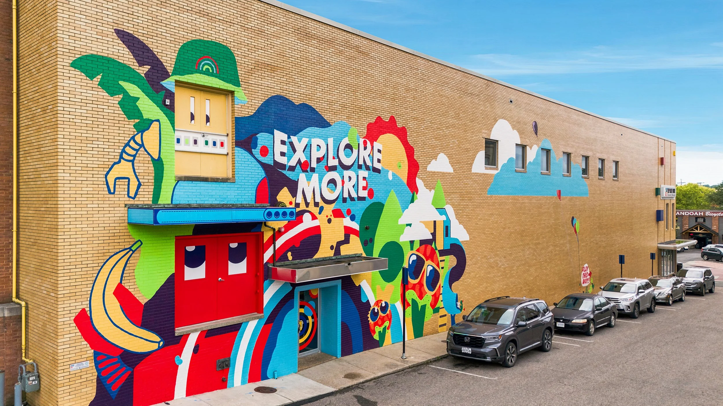 Colorful mural on a brick building wall with the phrase 'Explore More,' featuring abstract shapes, clouds, a rainbow, mountains, and various vibrant illustrations.
