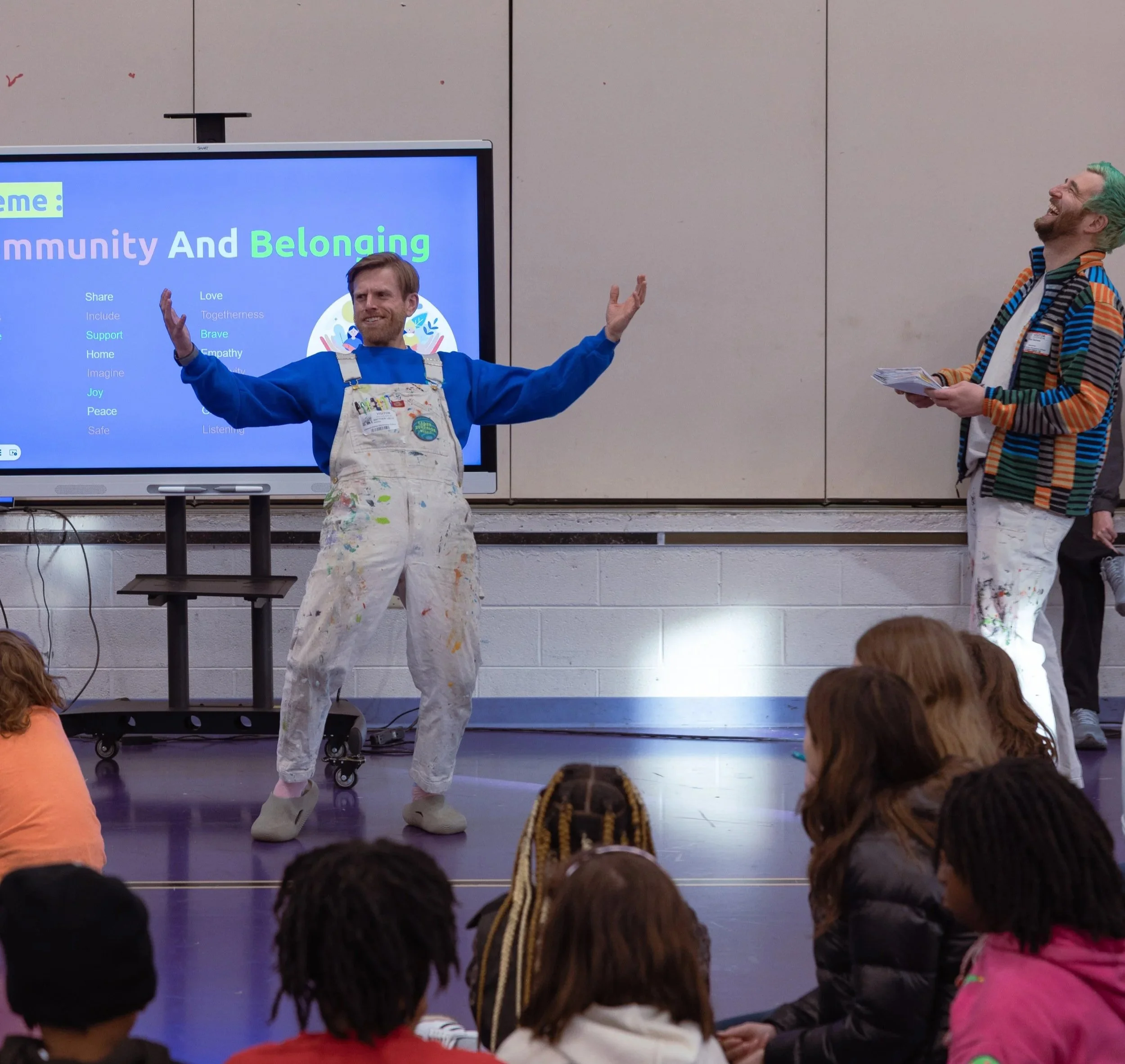 Two men are giving a presentation or performance in front of a seated audience of children. The man on the left is wearing a colorful apron and a blue shirt, gesturing with his hands while smiling. The man on the right has green hair, a striped jacket, and is holding notes, smiling as he looks at the other man. Behind them is a screen displaying the words "Community And Belonging" with a list of related words on a blue background.