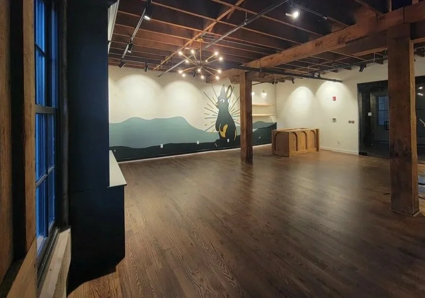 Empty interior space with wooden floors and exposed wooden beams on the ceiling. A large mural of a black rabbit with yellow accents and radiating lines is on the back wall. There is a chandelier hanging from the ceiling and a small wooden structure near the backwall. Large windows on the left side letting in natural light.