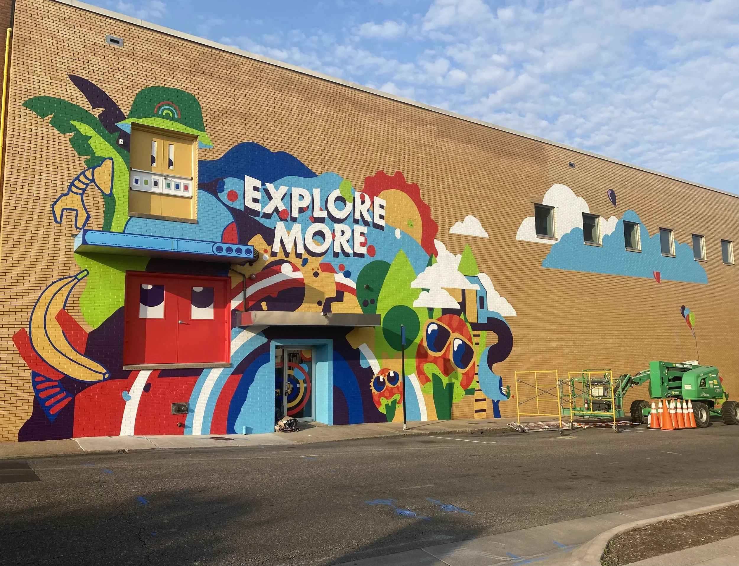 Colorful mural on the side of a brick building with the phrase "Explore More" amid illustrations of nature, animals, and abstract patterns. There are construction cones and equipment in front of the building.