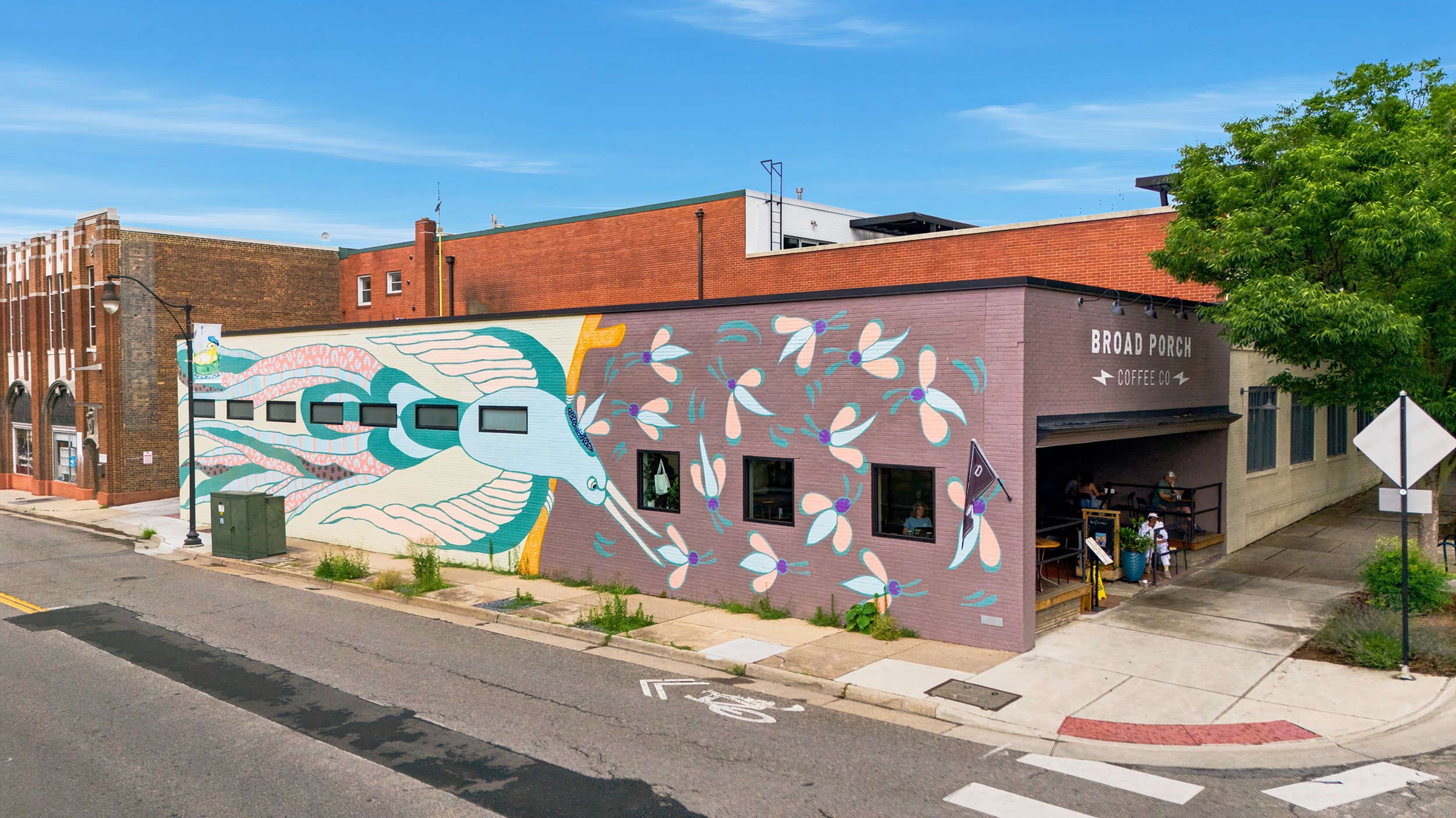 Street view of a brick building with a mural, a tree, a sidewalk, a crosswalk, and a sign for Broad Porch Coffee Co.