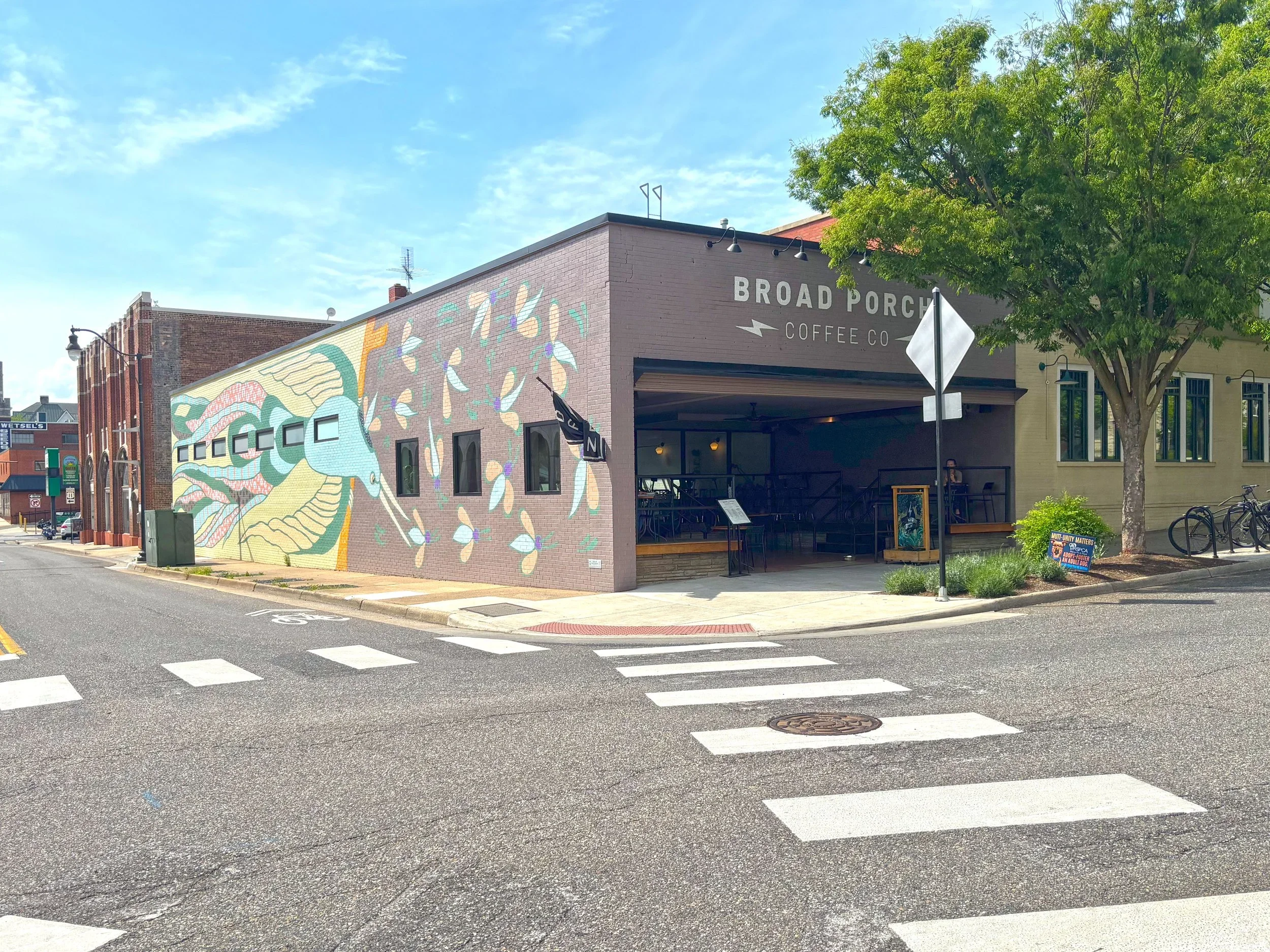 Street view of a building with a mural on the side, sign reading 'BROAD PORCH COFFEE CO,' outdoor seating, tree, and a pedestrian crosswalk.