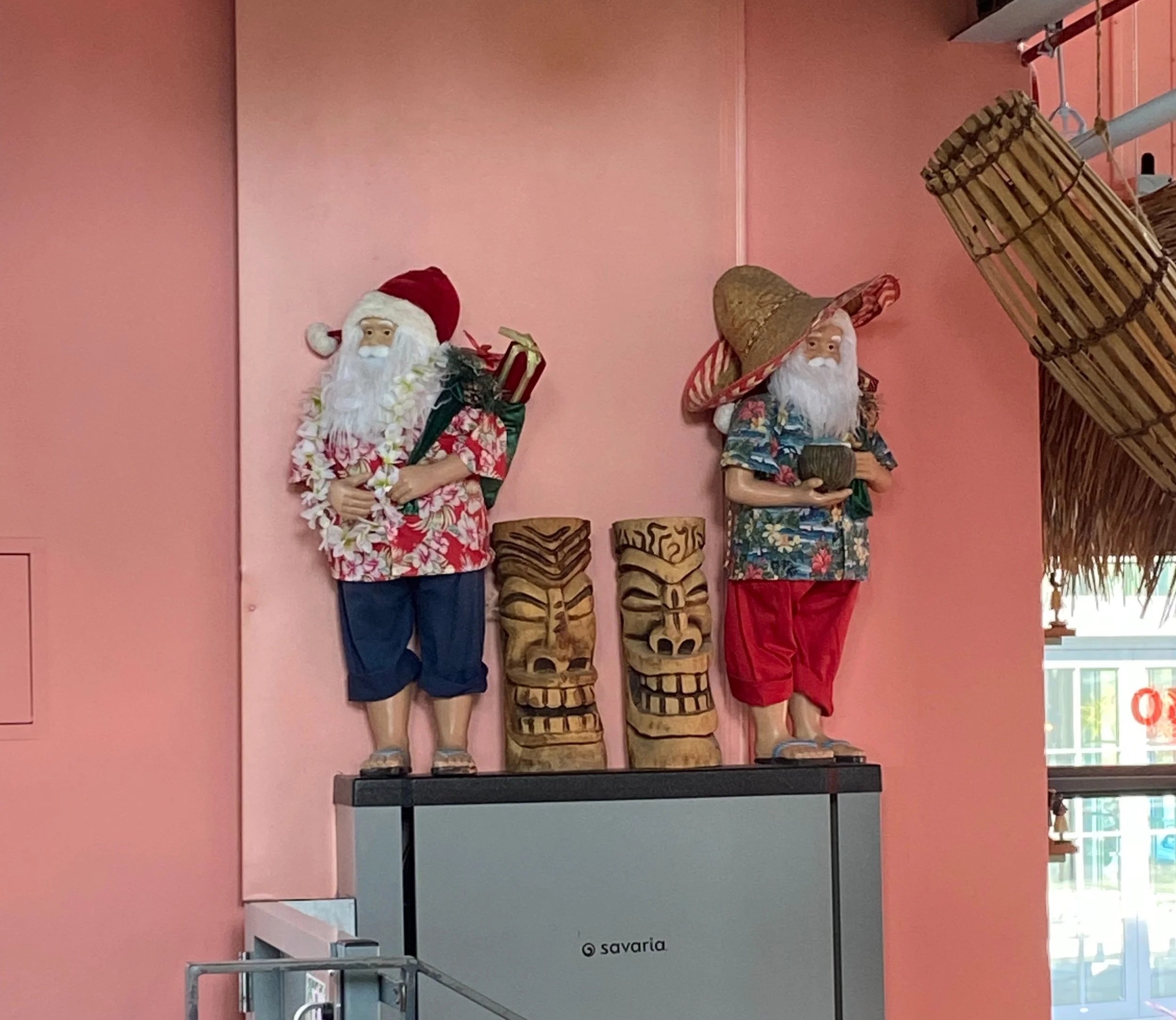 Decorative Santa Claus and pirate Santa figurines on a pink wall. Santa is dressed in a Hawaiian shirt with a floral lei, holding a gift, and wearing a red hat. Pirate Santa is wearing a tropical shirt, red pants, and a large straw hat, holding a coconut.
