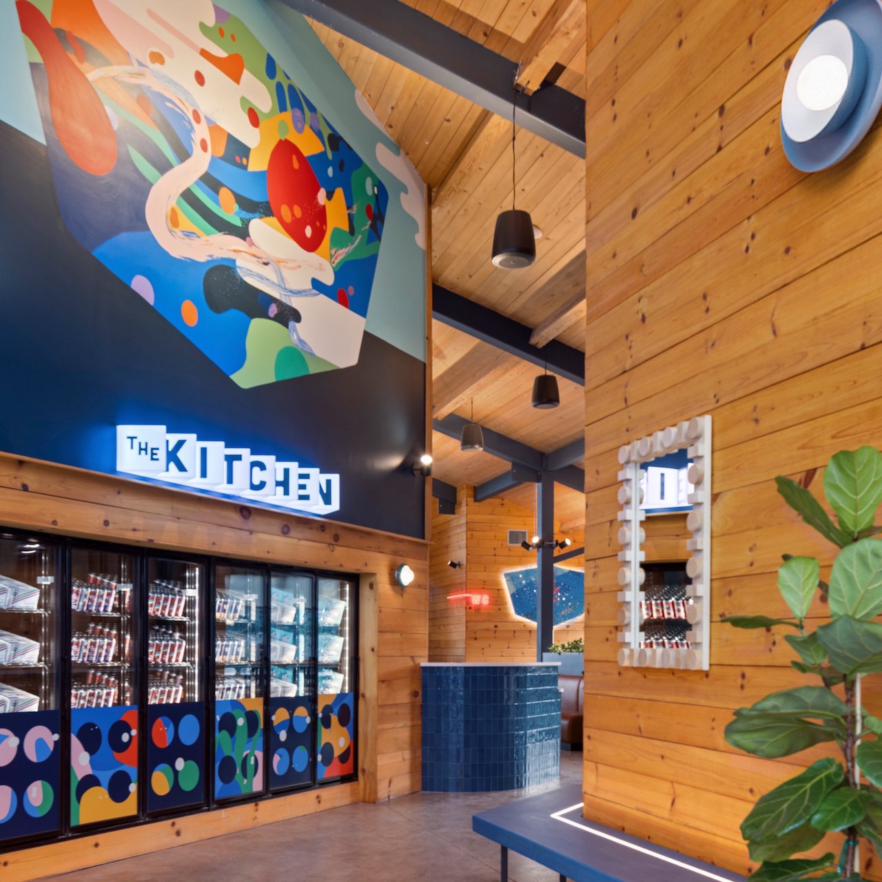 Interior of a restaurant called The Kitchen with wooden walls, a large colorful abstract mural, a mirror, and a refrigerator with drinks. There are black pendant lights hanging from the wooden ceiling and some decorative plants.