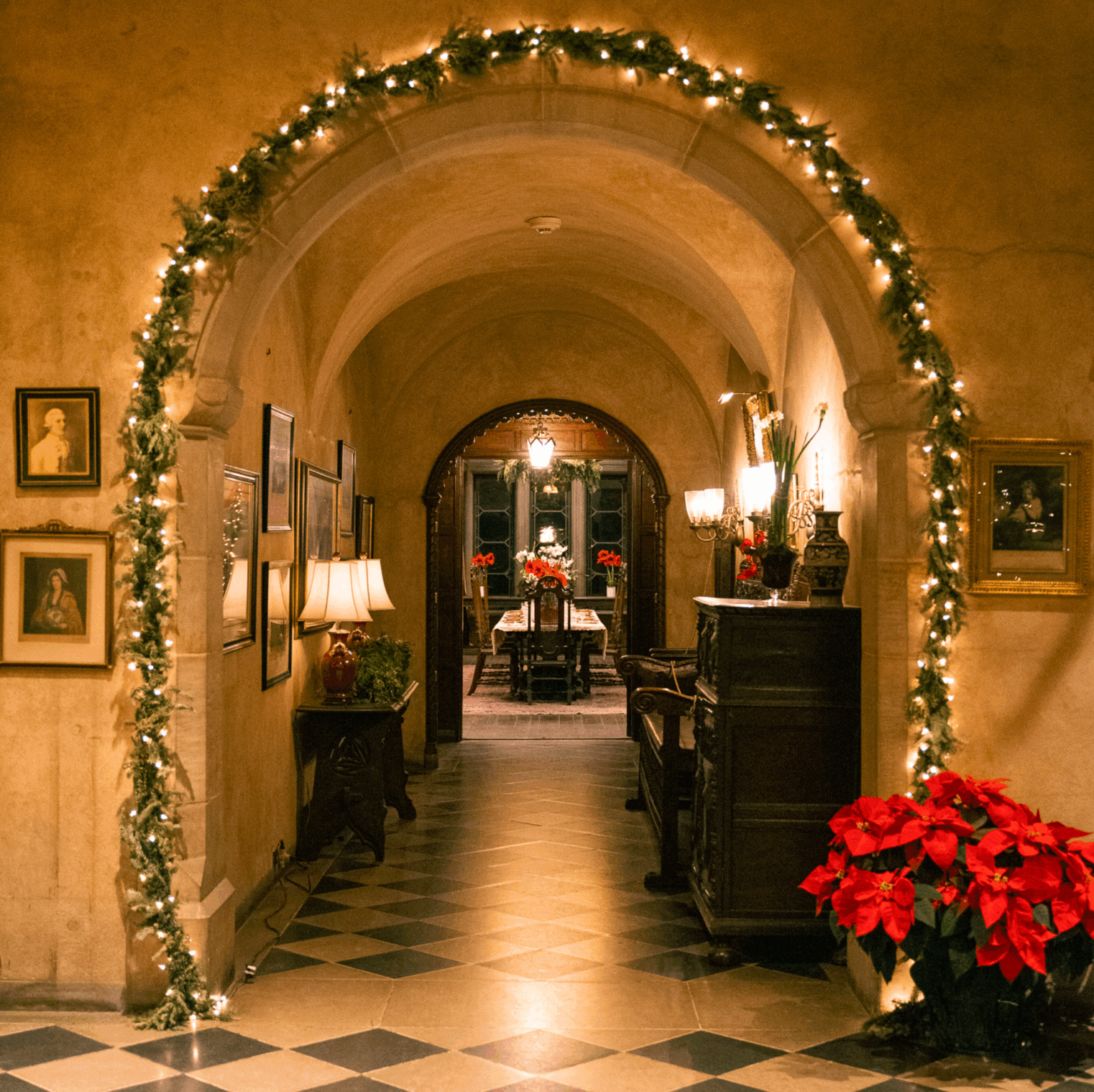 A warmly lit hallway in Coe Hall decorated with Christmas lights and poinsettias, leading to a dining room with a chandelier and festive decorations.