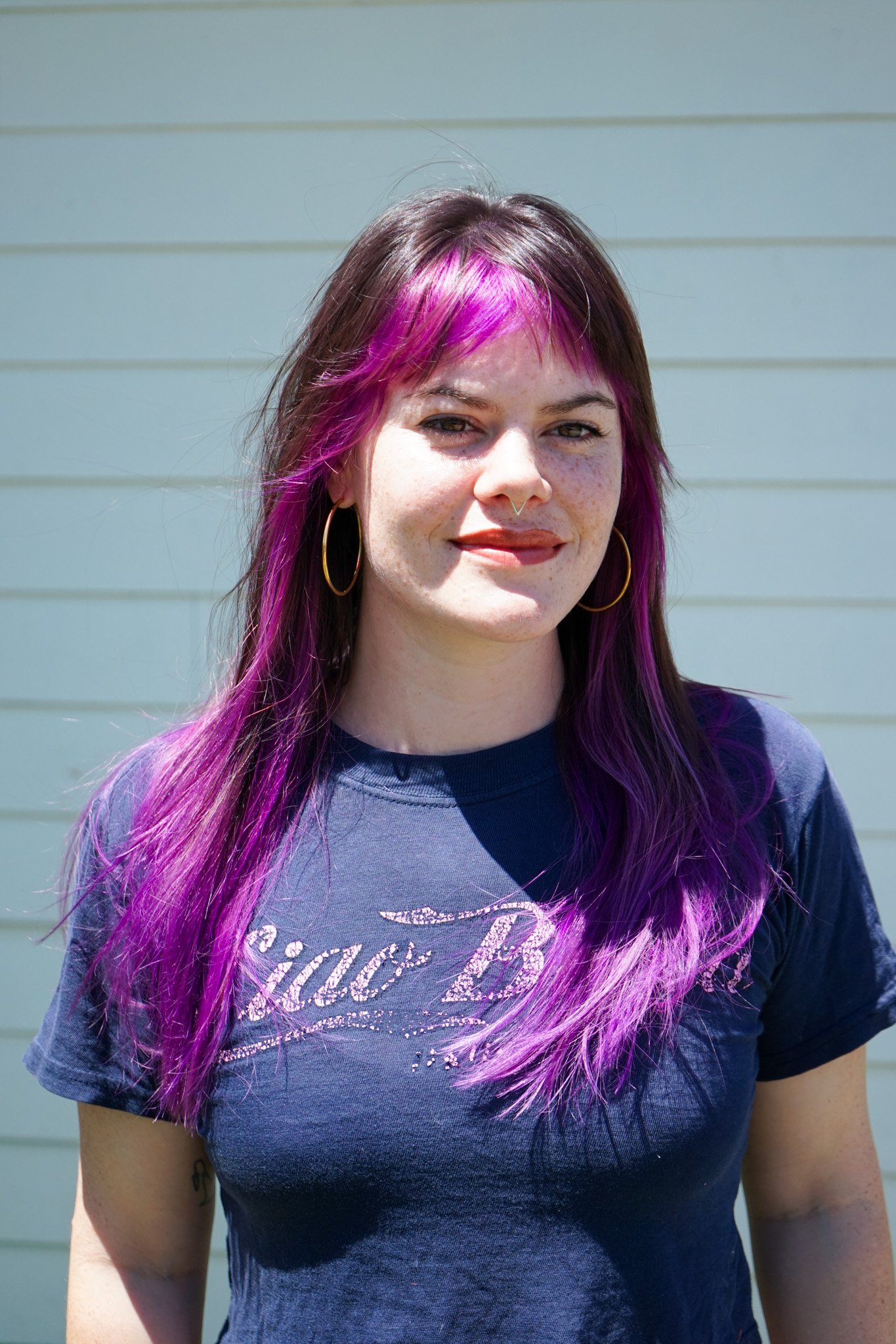 A young woman with shoulder-length brown hair with purple highlights, wearing gold hoop earrings and a navy blue T-shirt, standing outdoors in front of a light-colored, horizontal siding wall. She has a slight smile and is squinting slightly in the sunlight.