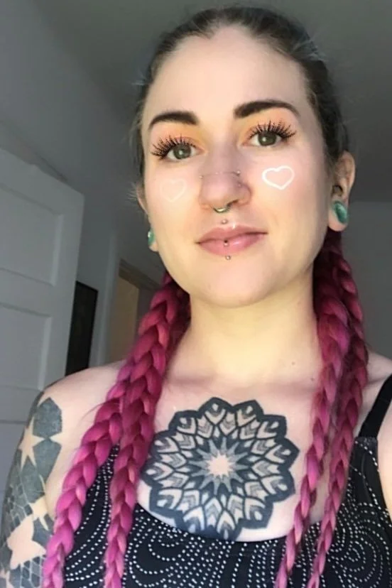 A young woman with pink braided hair, tattoos on her chest and arms, and makeup including labeled hearts and detailed eye makeup. She is smiling slightly, indoors.