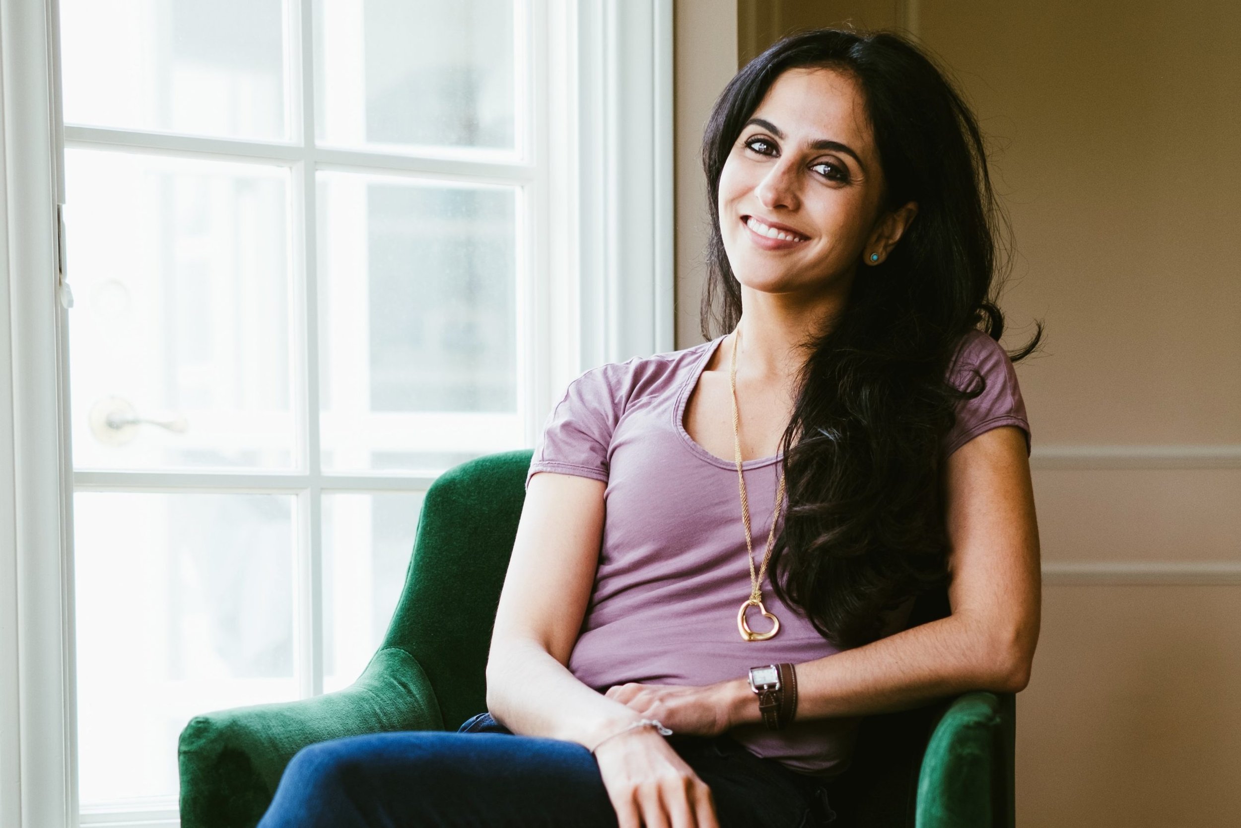 Ranavat founder Michelle Ranavat wearing a purple t-shirt, jeans, and gold heart necklace while sitting on a green velvet chair