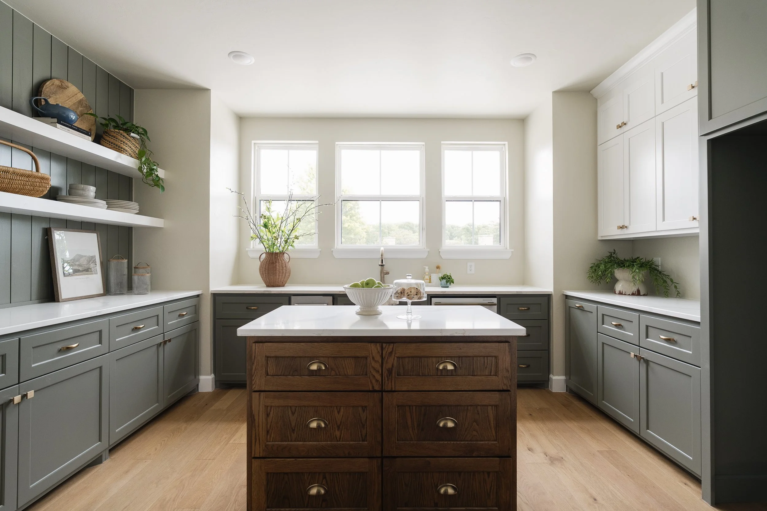 A modern kitchen with gray cabinets, white countertops, a black faucet, a window, and decorative items like a framed picture, a plant, and a basket.