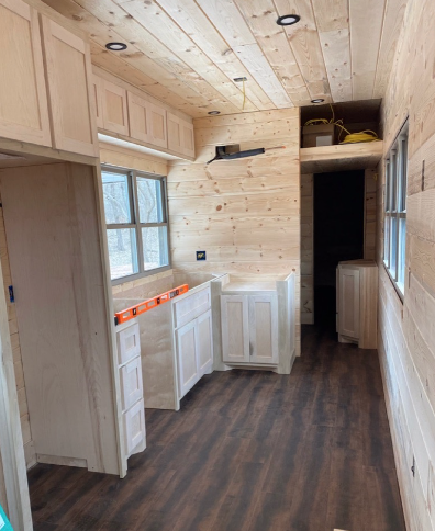 Interior of a partially built room with wooden walls and ceiling, with kitchen cabinets installed under windows and electrical wiring visible, and dark hardwood flooring.
