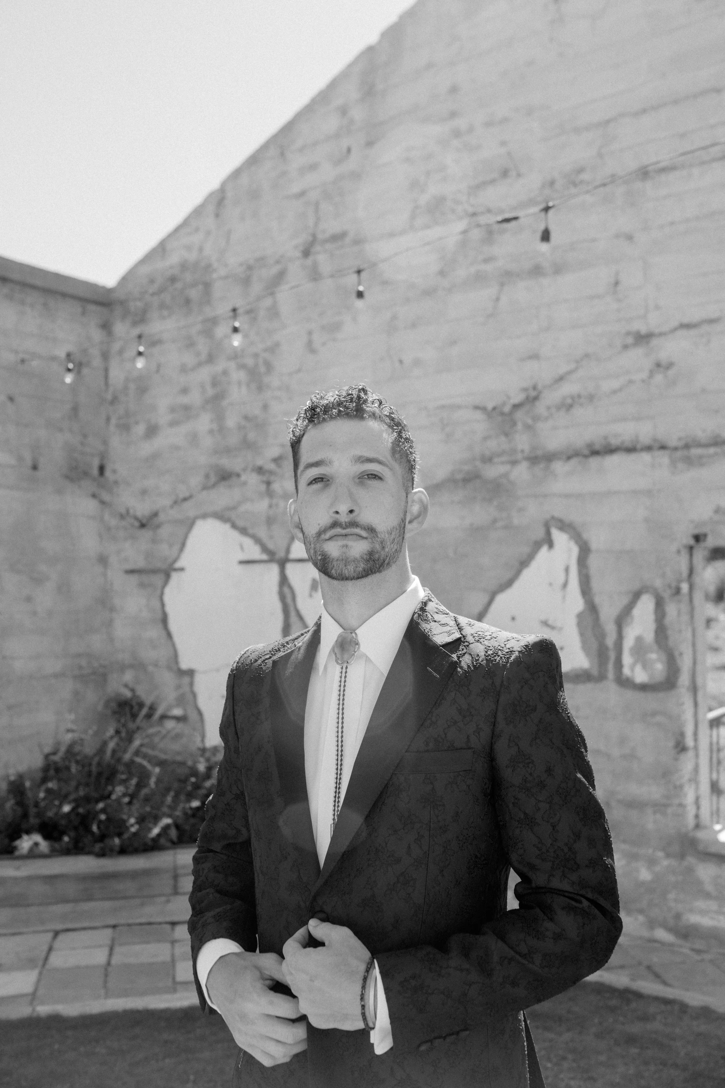 Man in formal suit standing in front of an old textured wall with string lights, black and white photo.